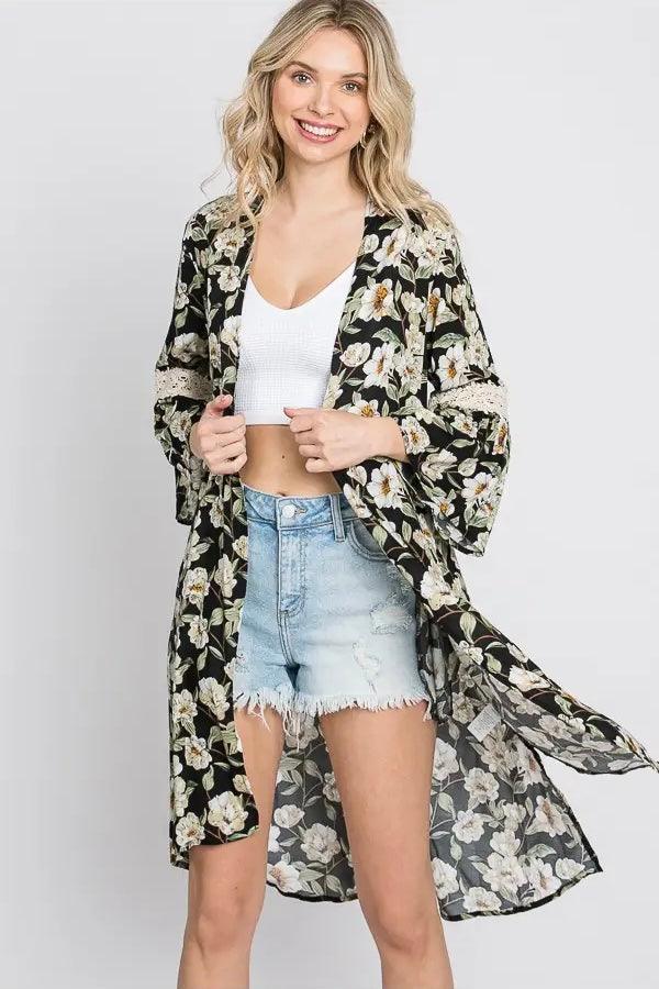 Smiling woman wearing black floral print 3/4 bell sleeve duster over white crop top and distressed denim shorts