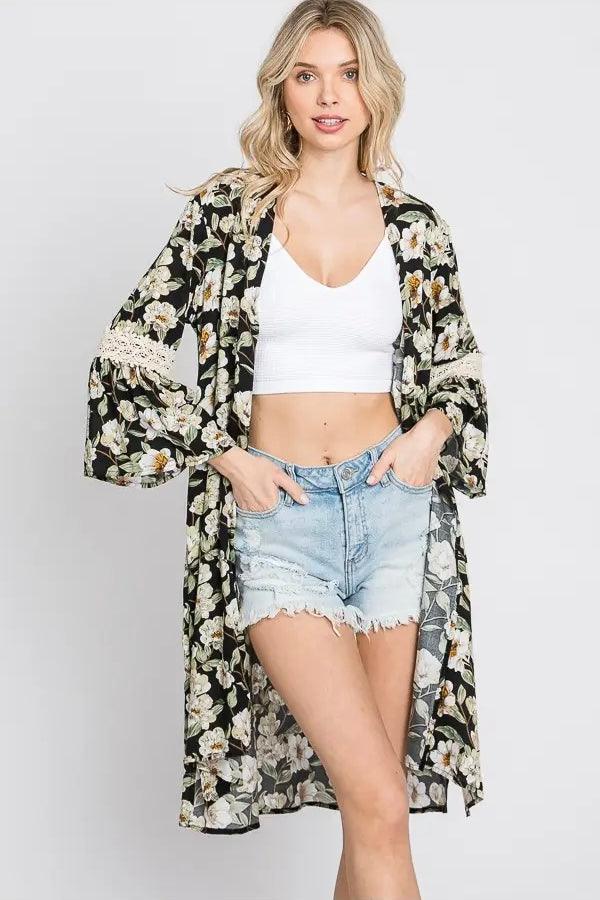 Blonde woman wearing floral print 3/4 bell sleeve duster over white crop top and light denim shorts against gray background