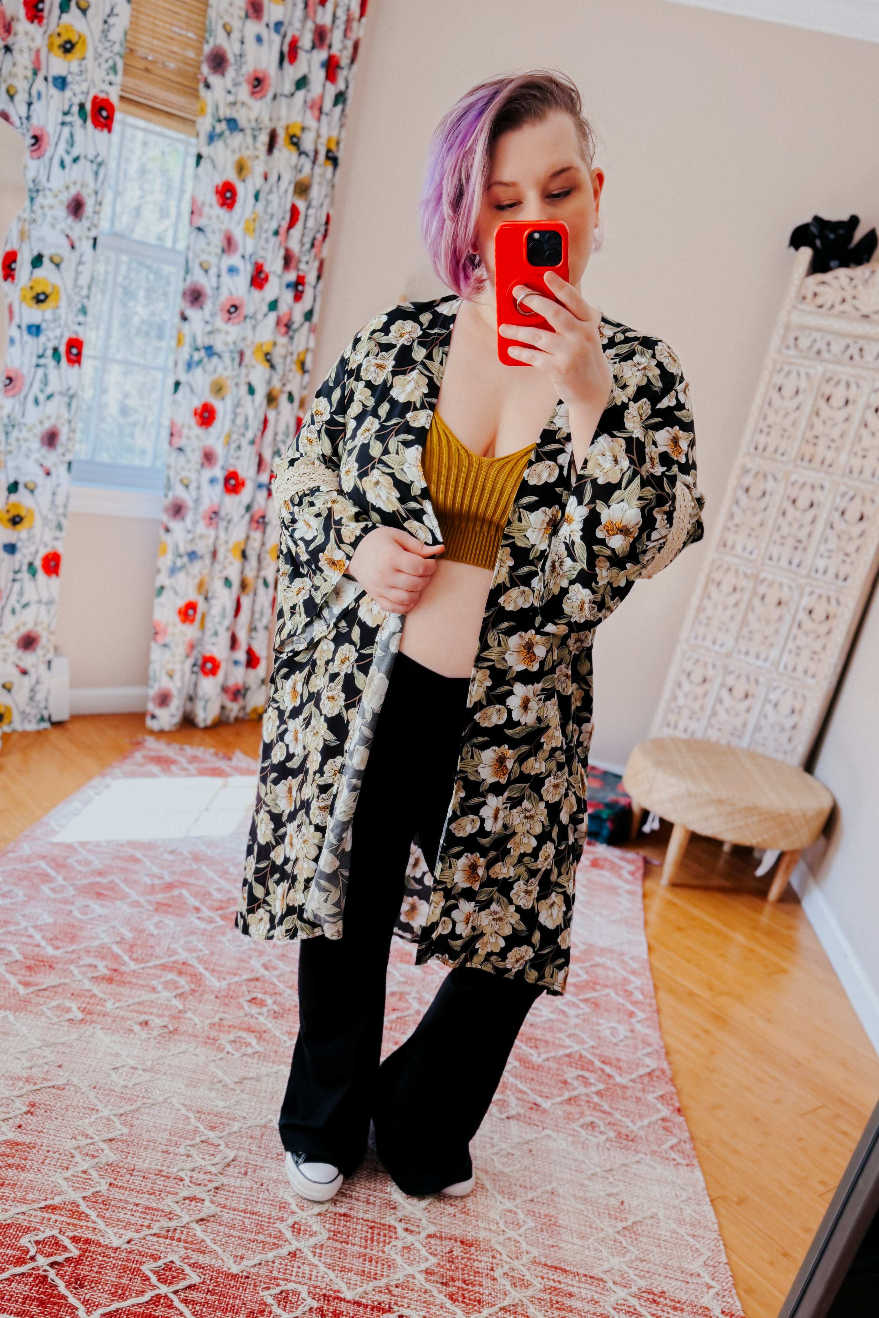 Woman with short purple hair wearing floral print 3/4 sleeve duster over yellow top and black pants taking mirror selfie indoors