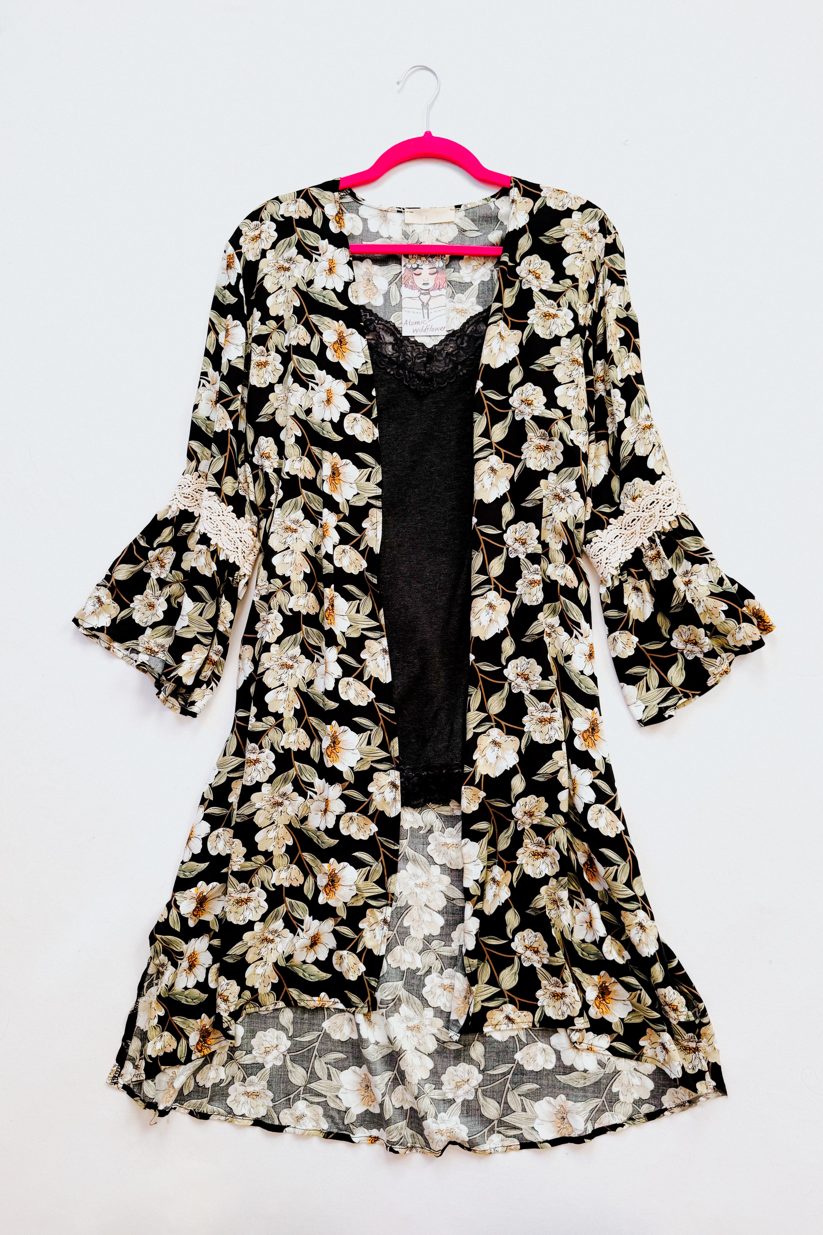 Black floral duster with bell sleeves and lace trim layered over black lace-trimmed camisole on pink hanger against white background