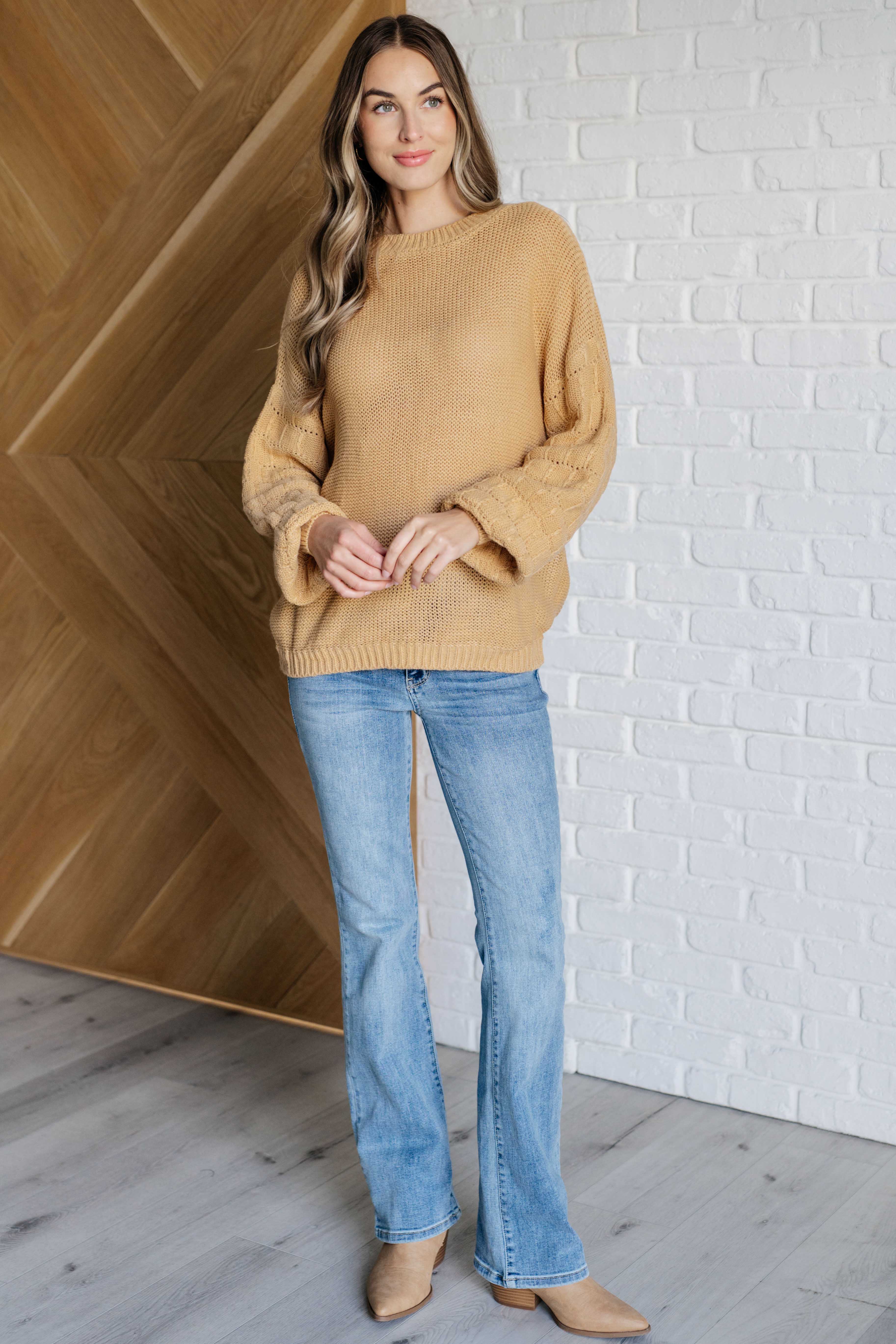 Woman wearing wheat-colored knit sweater with voluminous sleeves and light blue flared jeans standing indoors