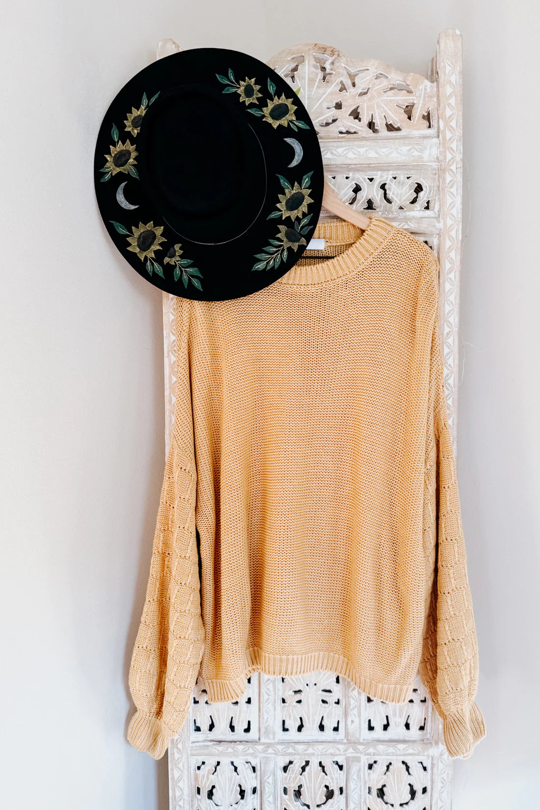 Wheat-colored bubble sleeve sweater with textured knit hanging on carved white wooden panel with black floral hat