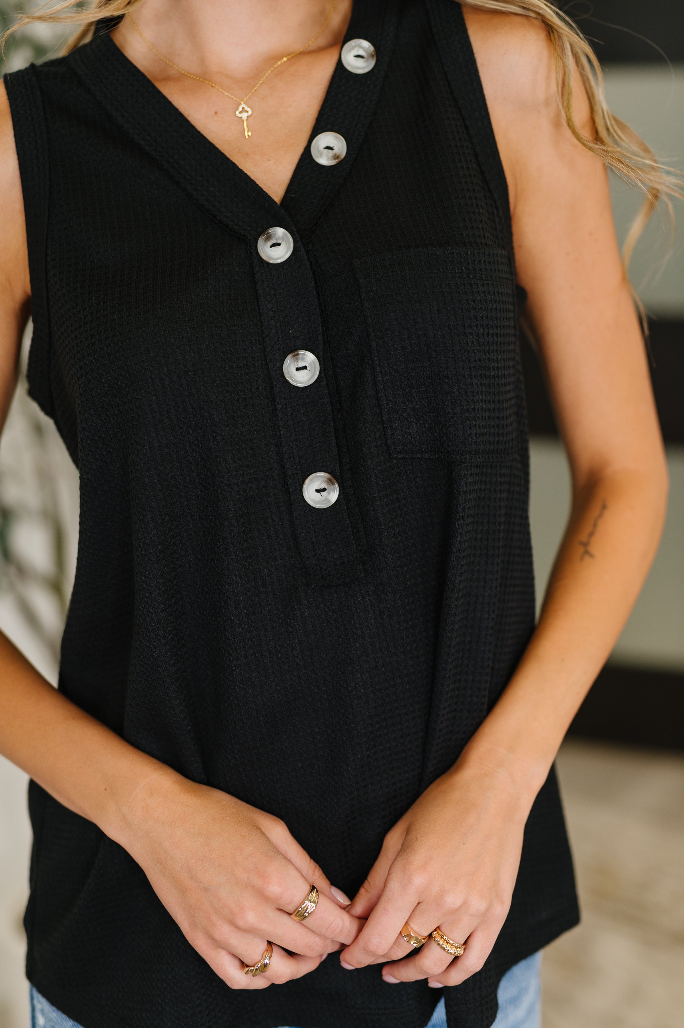 Woman wearing black sleeveless top with button details and small chest pocket, styled with delicate jewelry and rings
