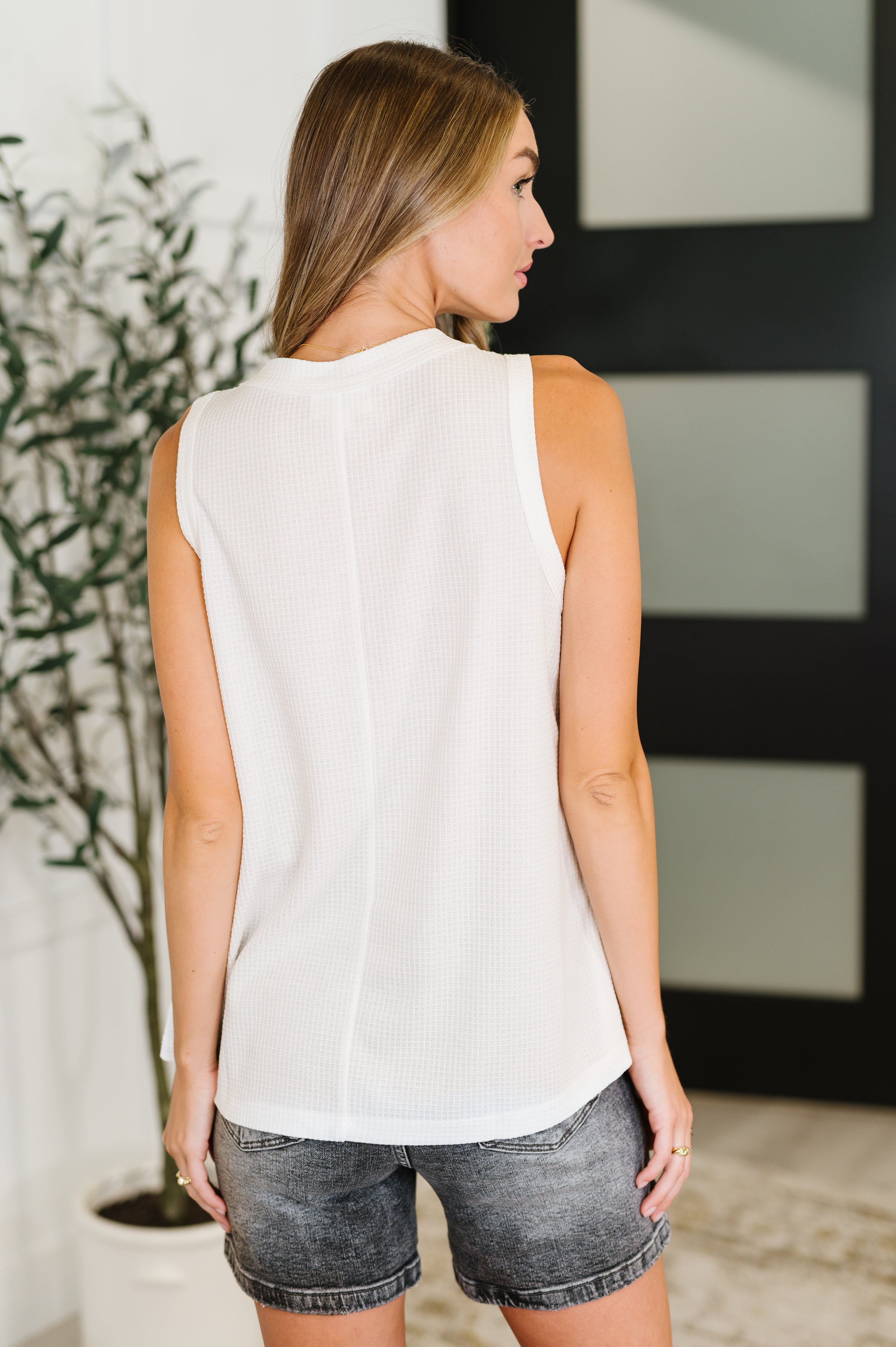 Woman wearing ivory sleeveless textured top with center seam paired with faded black denim shorts indoors
