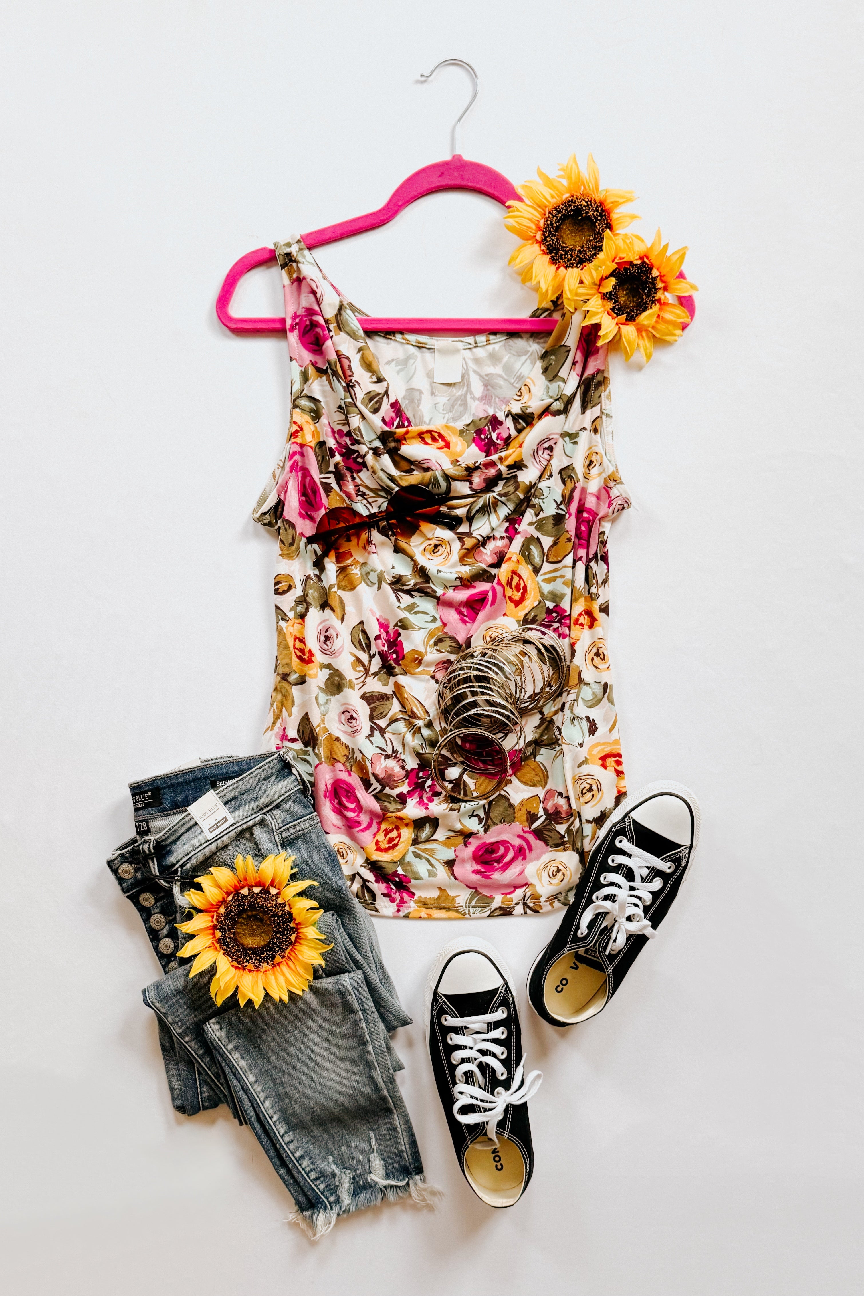 Floral cowl neck tank top on pink hanger with bracelets, distressed jeans, sunflower, and black sneakers on white background