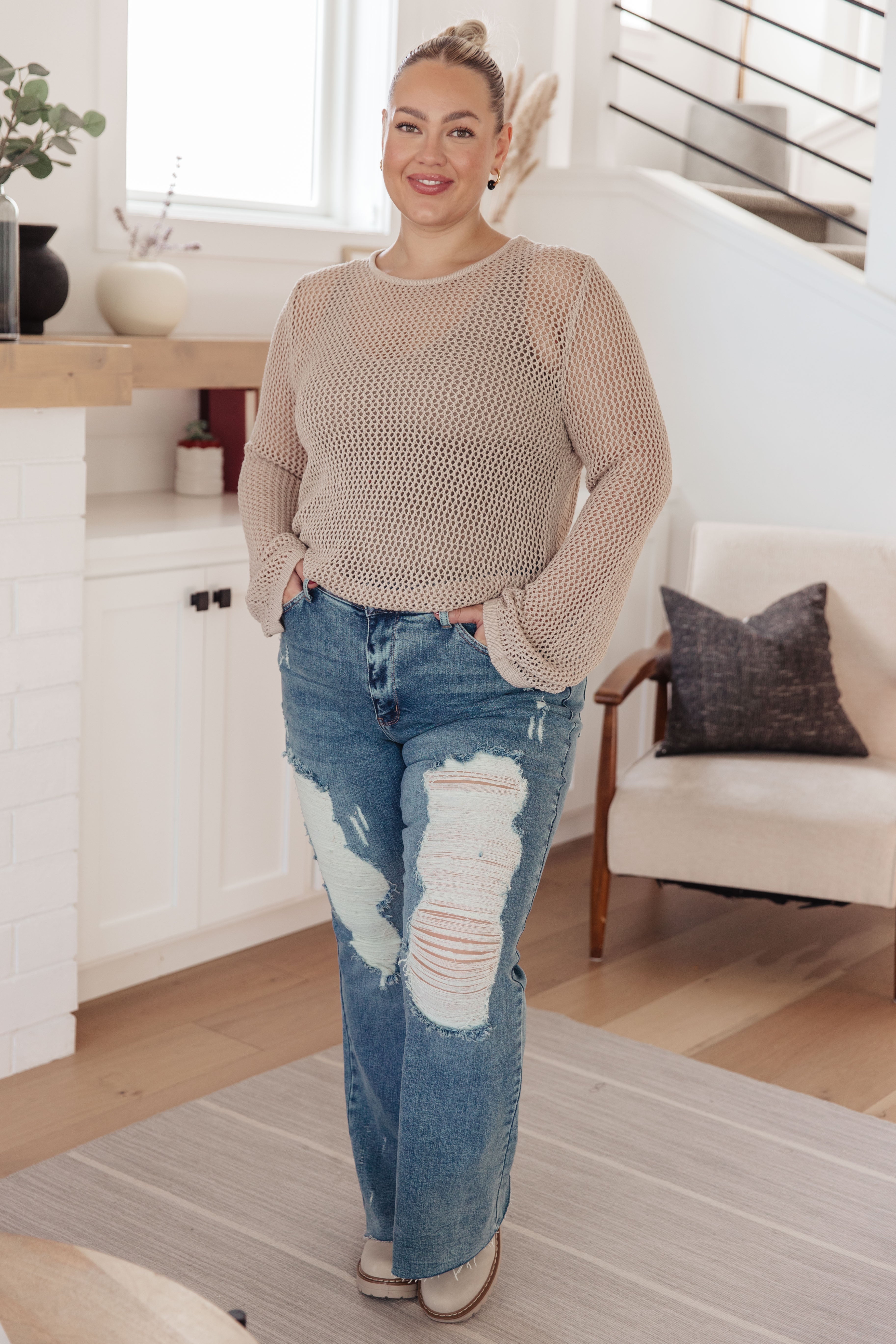 Woman wearing beige loose knit long sleeve top paired with distressed wide-leg jeans standing indoors smiling