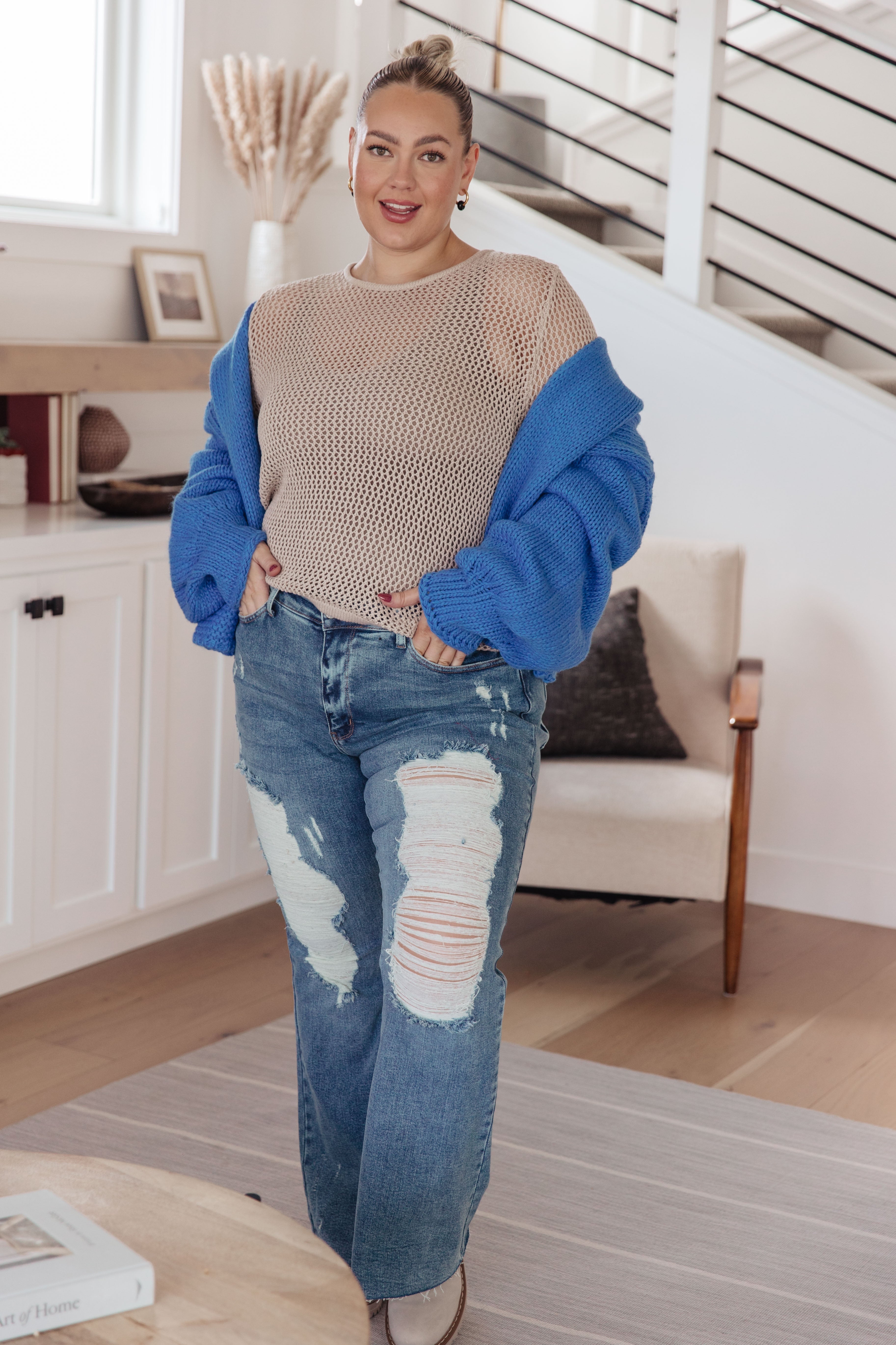 Woman wearing beige loose knit top with oversized blue cardigan and ripped jeans standing indoors in bright room