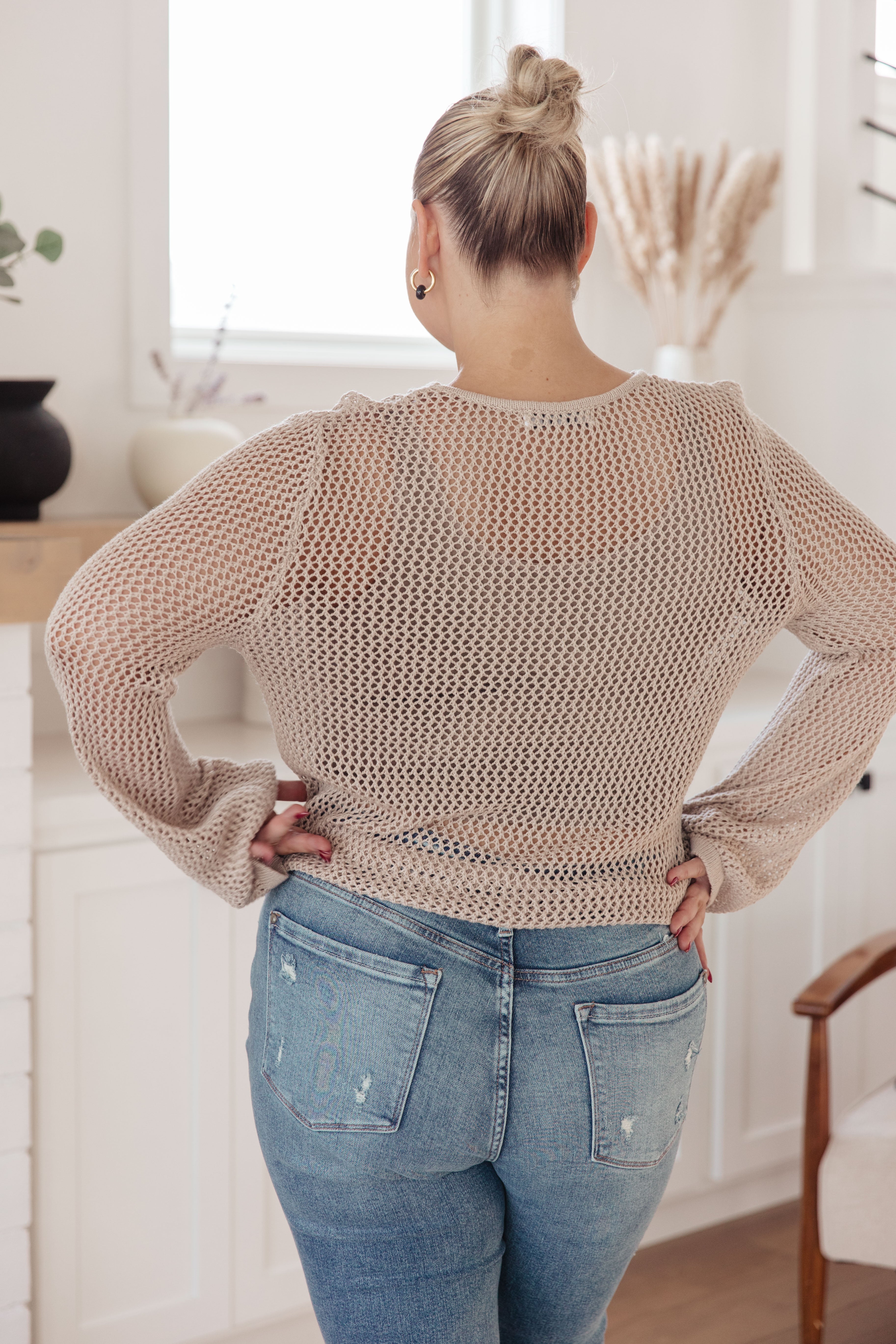 Woman wearing beige open knit loose long sleeve top with light distressed blue jeans standing indoors back view