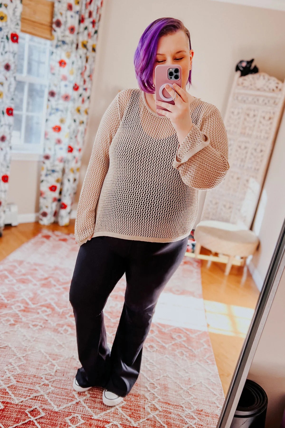 Woman with purple hair wearing loose beige knit long sleeve top and black pants taking mirror selfie indoors