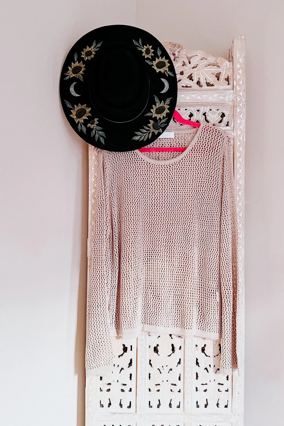 Light beige open knit long sleeve top hanging on carved white wooden panel with black floral embroidered hat