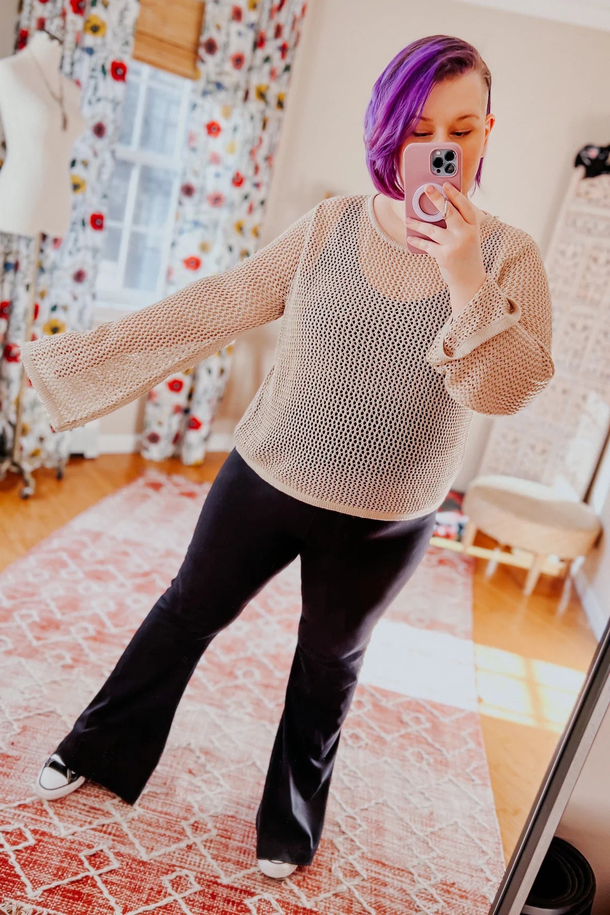 Woman with purple hair wearing loose beige knit top and black pants taking mirror selfie in bright room