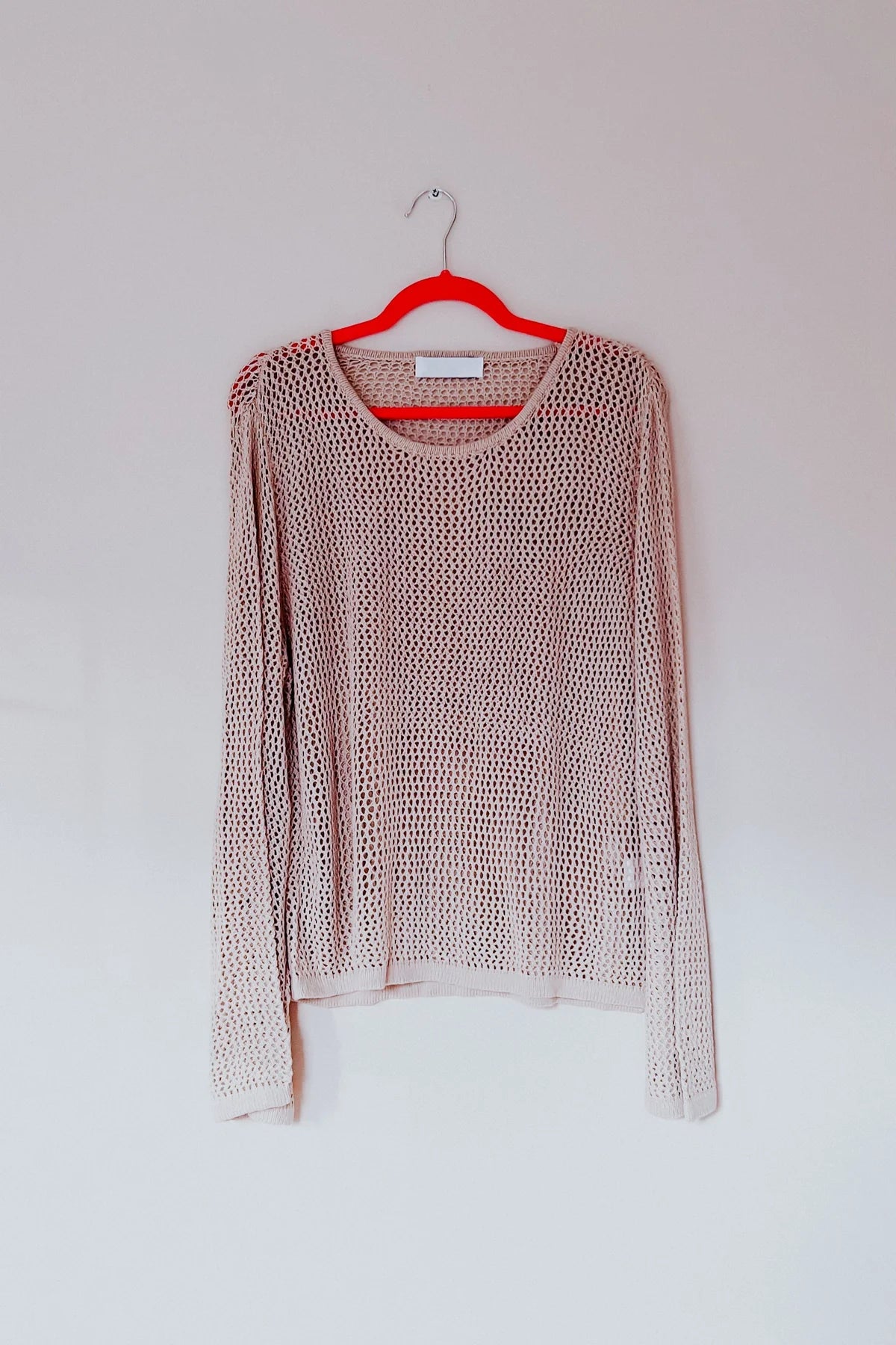 Light beige loose knit long sleeve top hanging on red hanger against plain wall