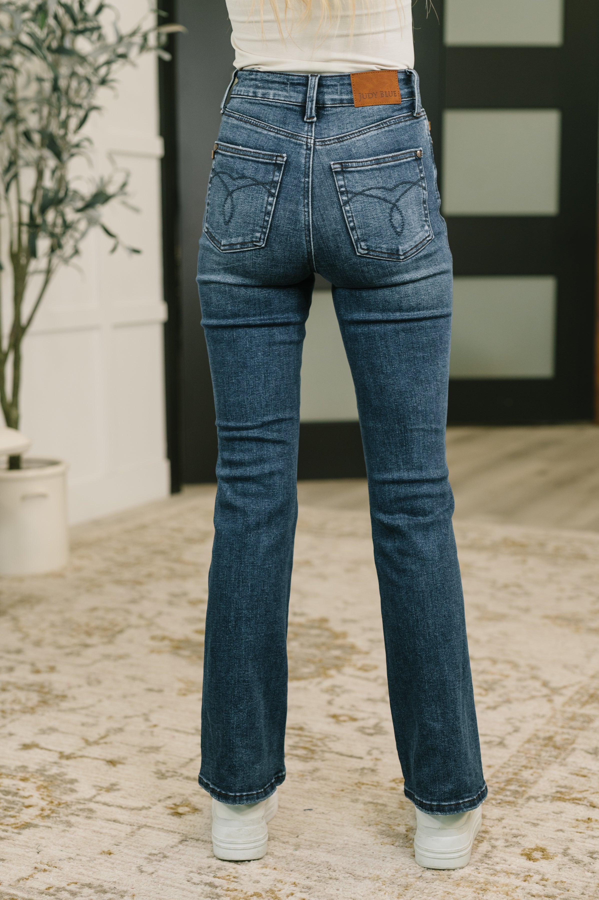 Back view of a woman in dark blue high waist bootcut jeans with back pockets and white sneakers indoors