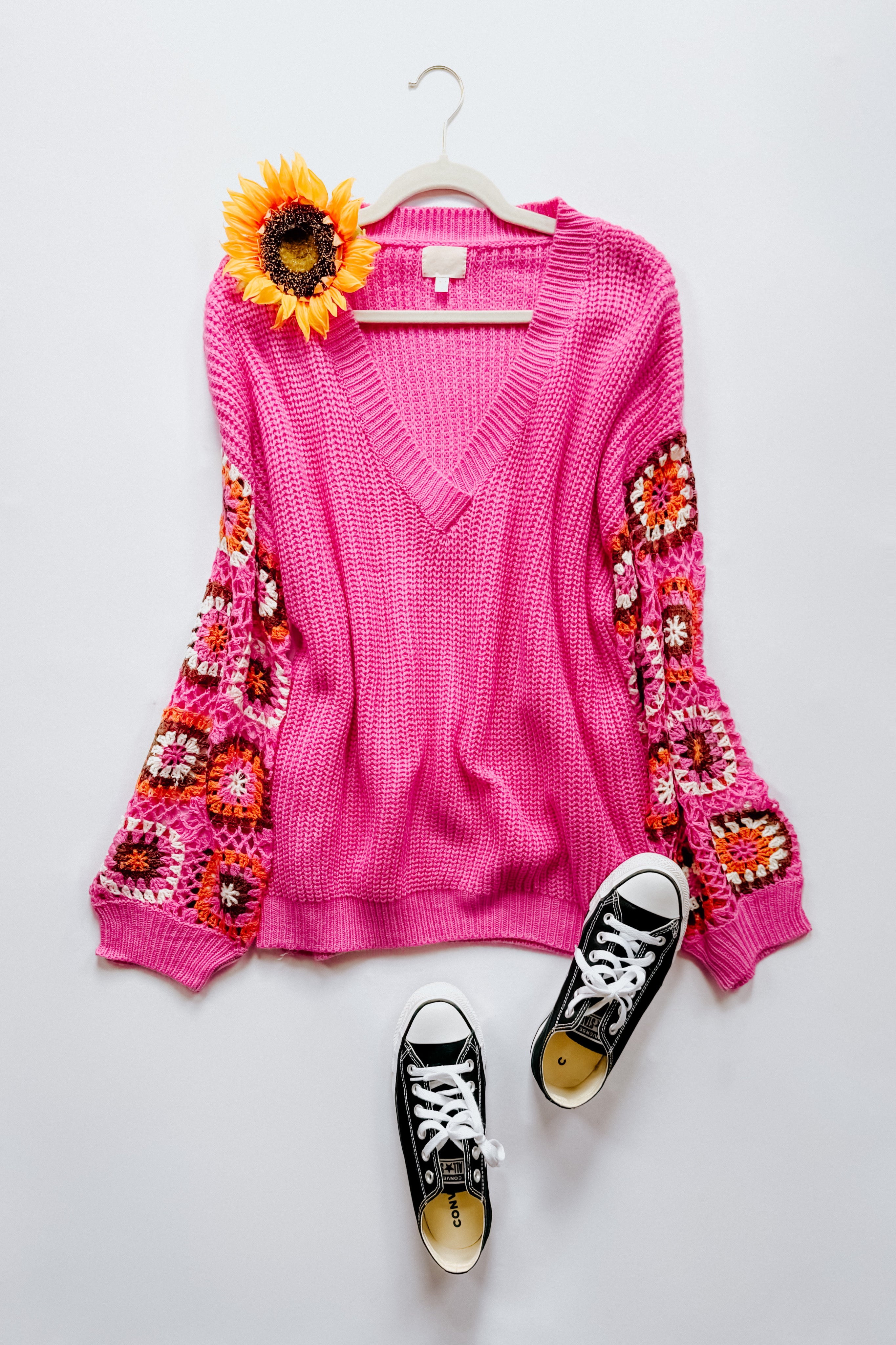 Bright pink V-neck knit sweater with multicolored crochet patchwork sleeves styled with black low-top sneakers and sunflower accessory