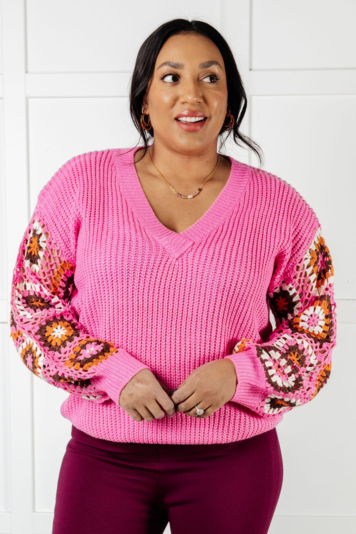Woman wearing bright pink chunky knit V-neck sweater with multicolored crochet granny square balloon sleeves and dark purple pants