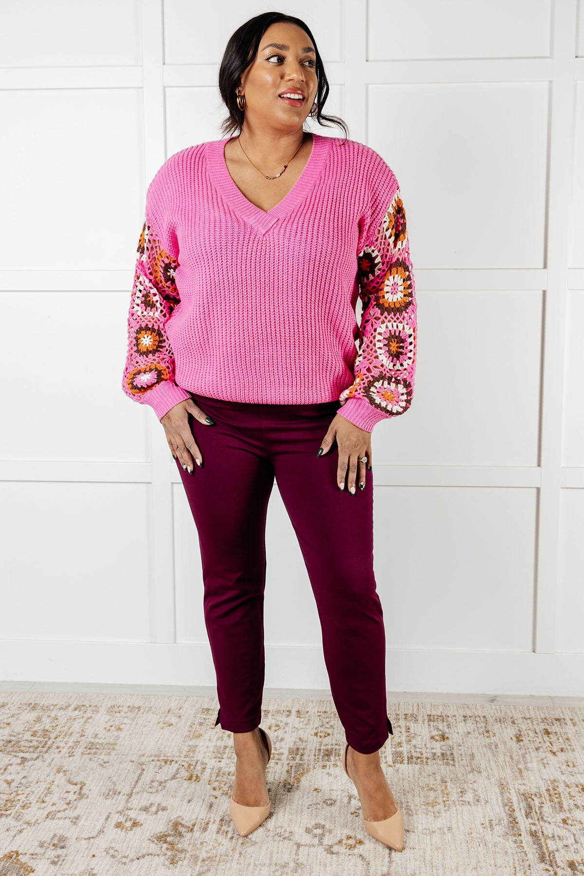 Woman wearing bright pink chunky V-neck sweater with colorful crochet granny square balloon sleeves and maroon pants