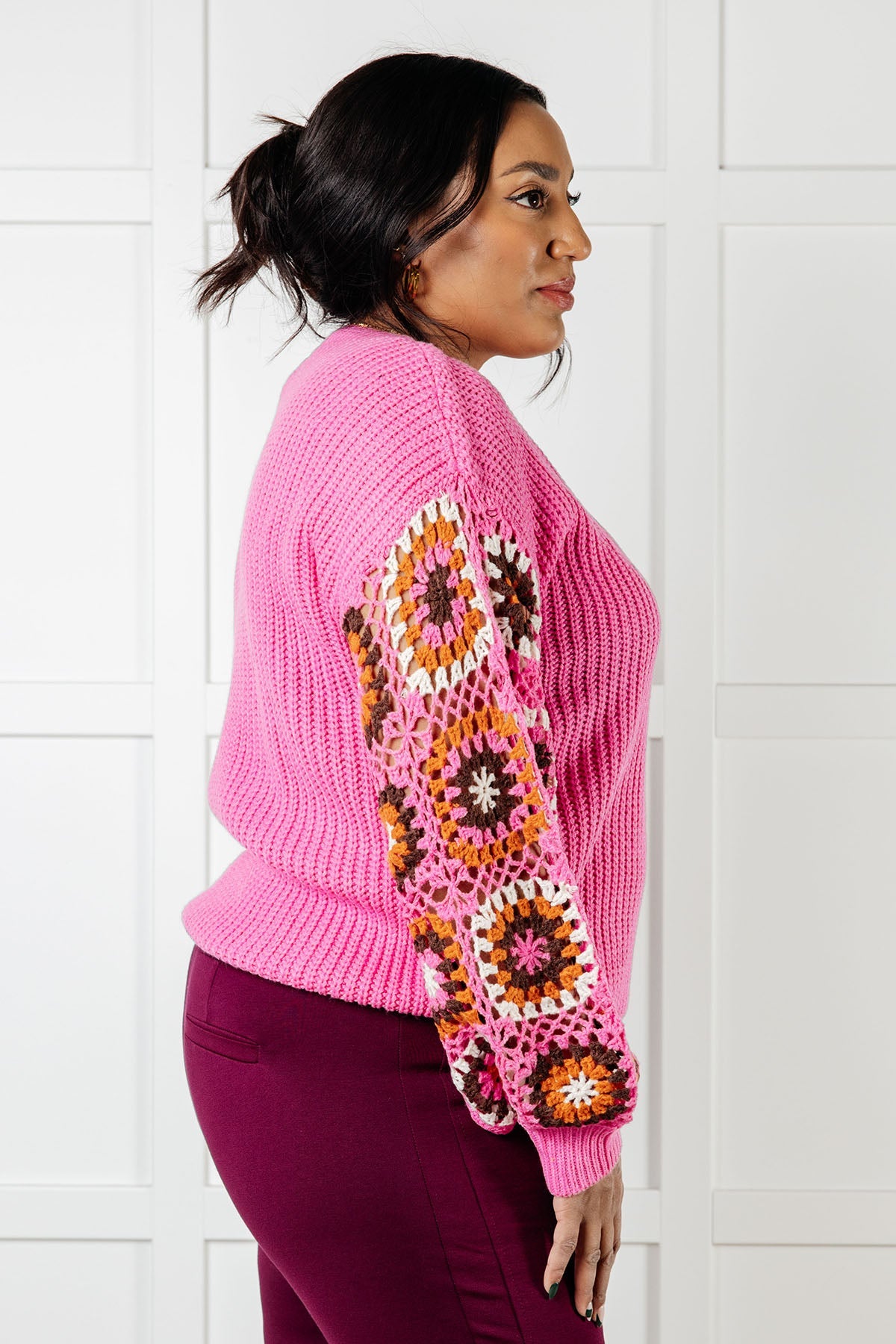 Side profile of woman wearing bright pink V-neck knit sweater with colorful crochet square patch sleeves and dark purple pants
