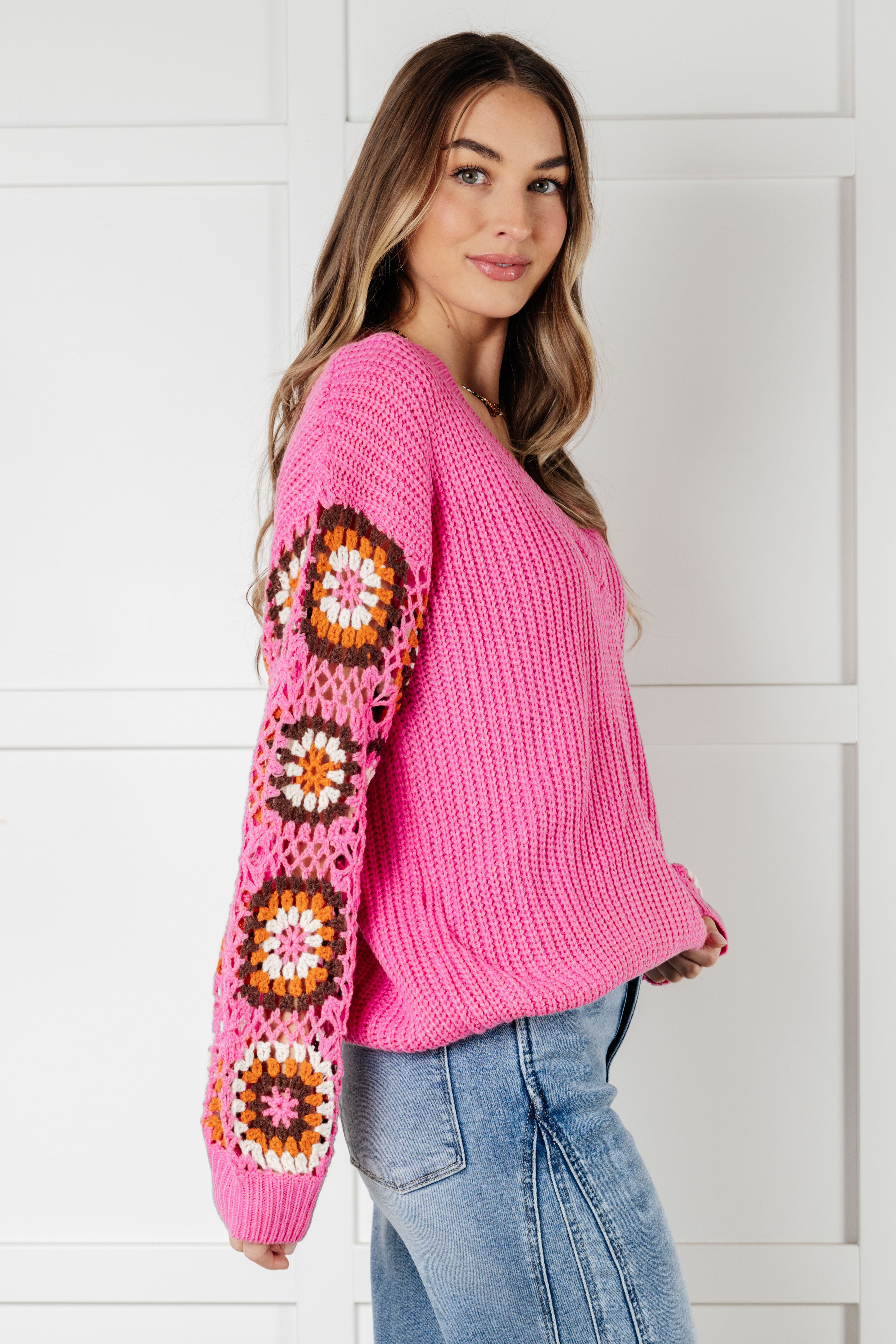 Side view of bright pink V-neck knit sweater with colorful crochet granny square sleeves worn with light wash jeans