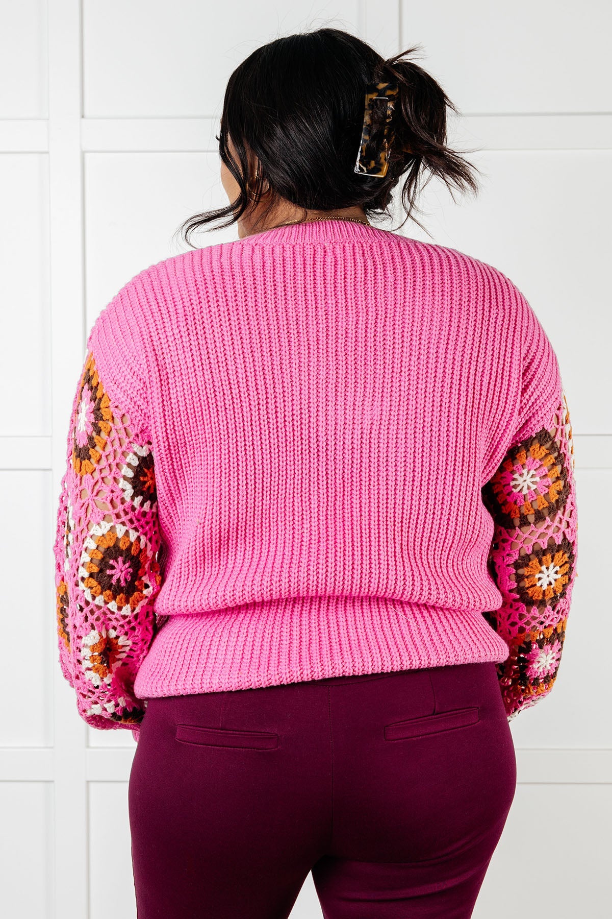 Back view of bright pink knit sweater with crochet floral sleeves worn with dark purple pants