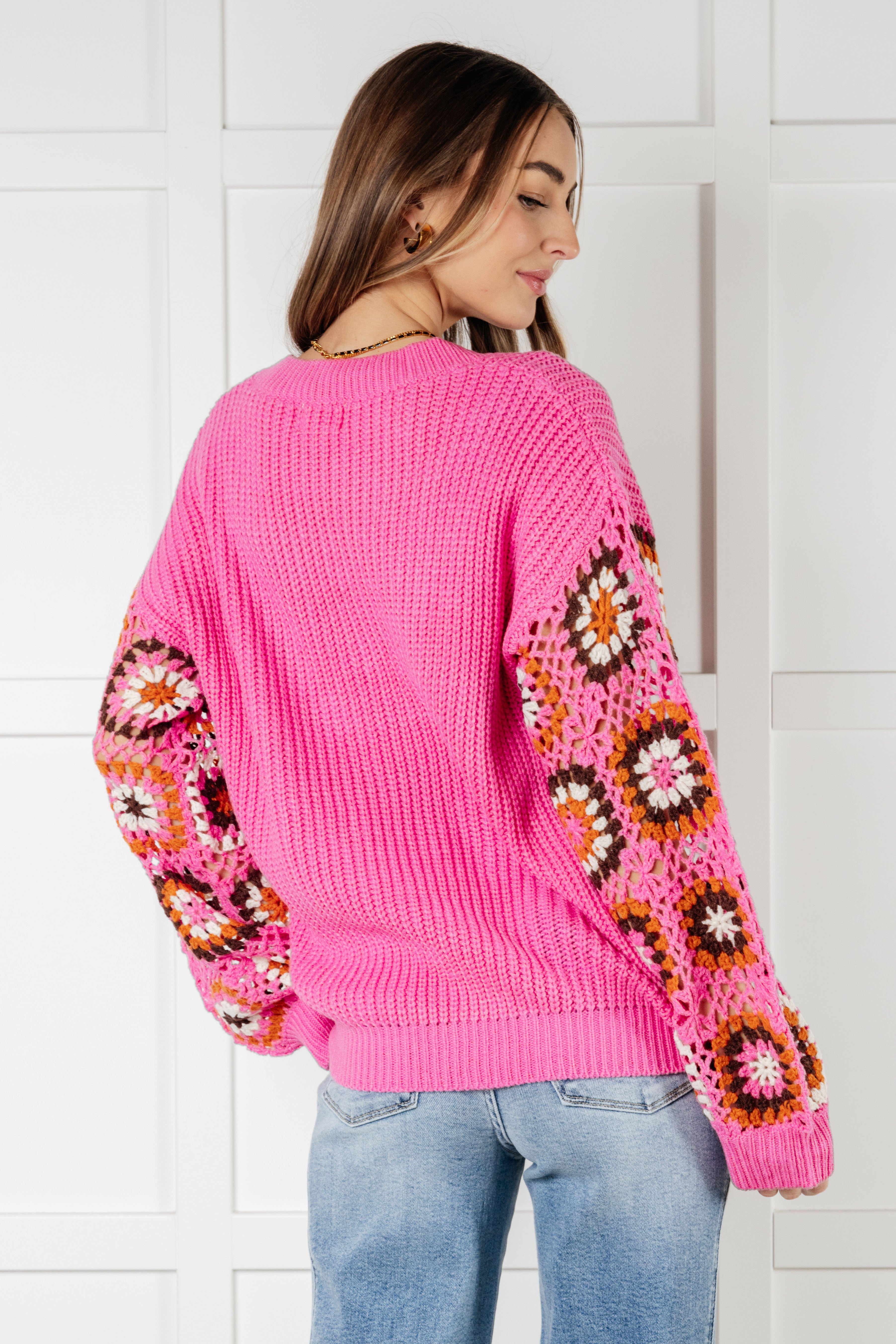 Woman modeling bright pink knit sweater with multicolored crochet floral pattern on sleeves and light blue jeans
