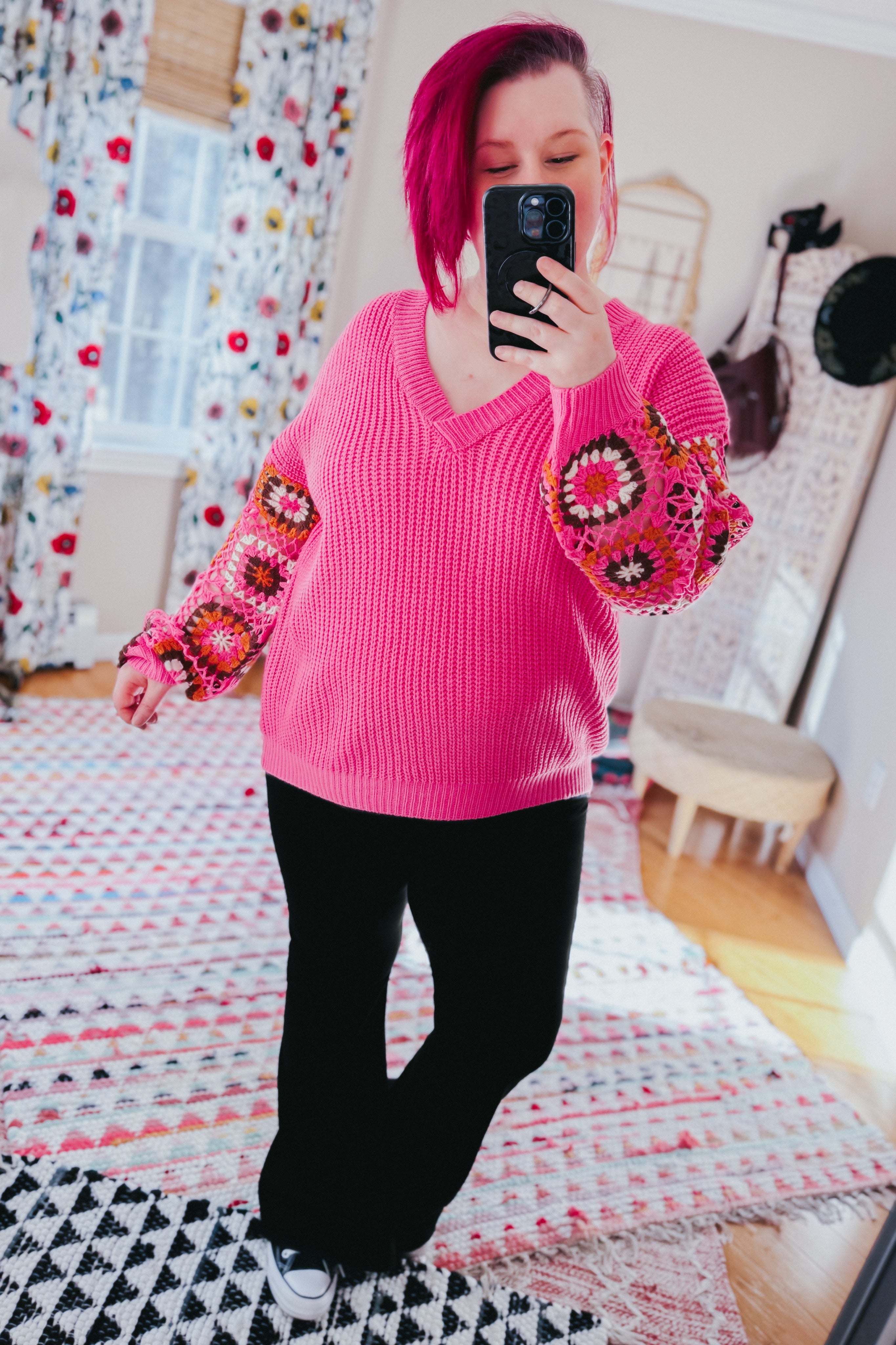 Woman taking mirror selfie wearing bright pink V-neck knit sweater with crochet patchwork sleeves and black pants indoors