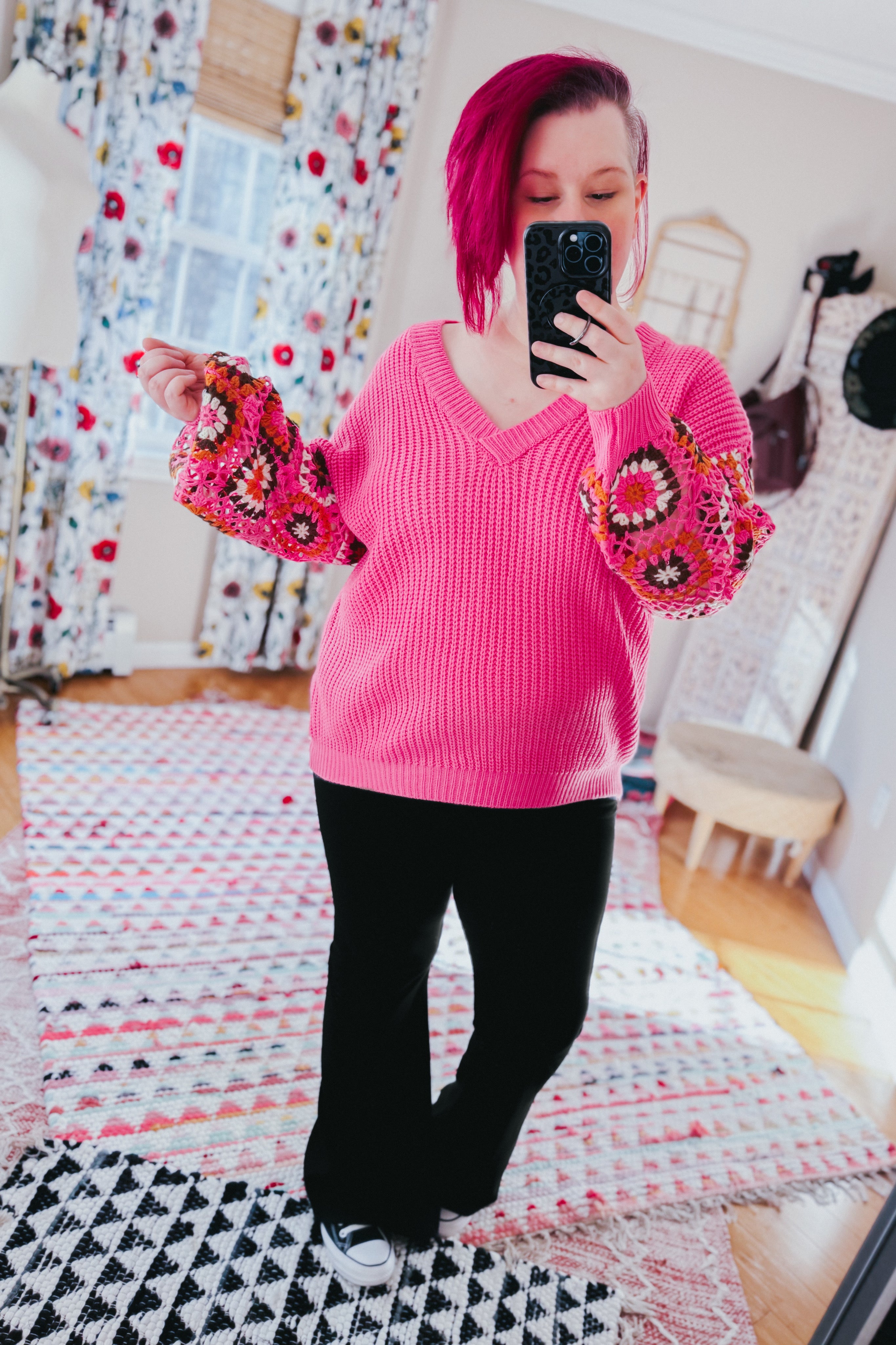 Woman with magenta hair wearing bright pink V-neck knit sweater with colorful crochet puff sleeves and black pants taking selfie indoors