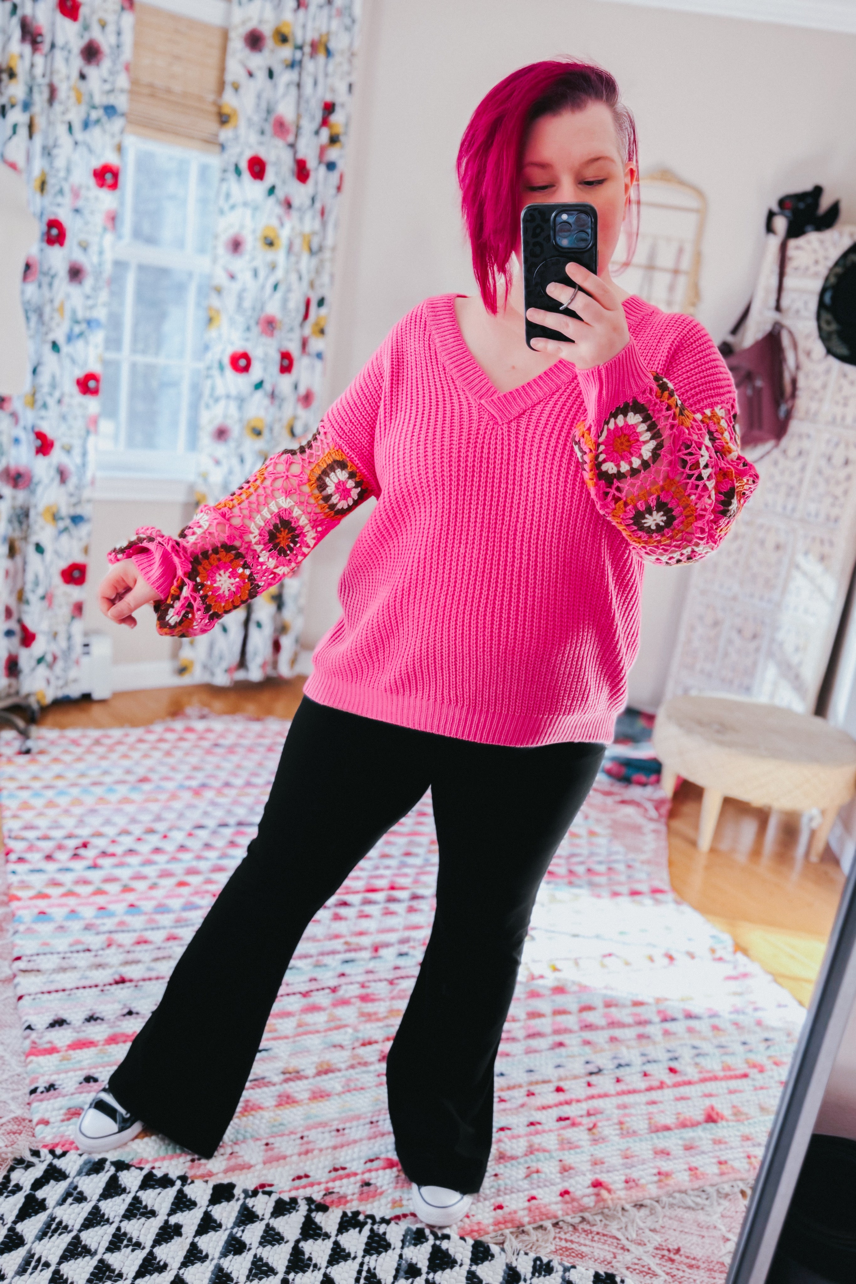 Woman wearing bright pink V-neck knit sweater with crochet patterned balloon sleeves and black pants indoors taking mirror selfie