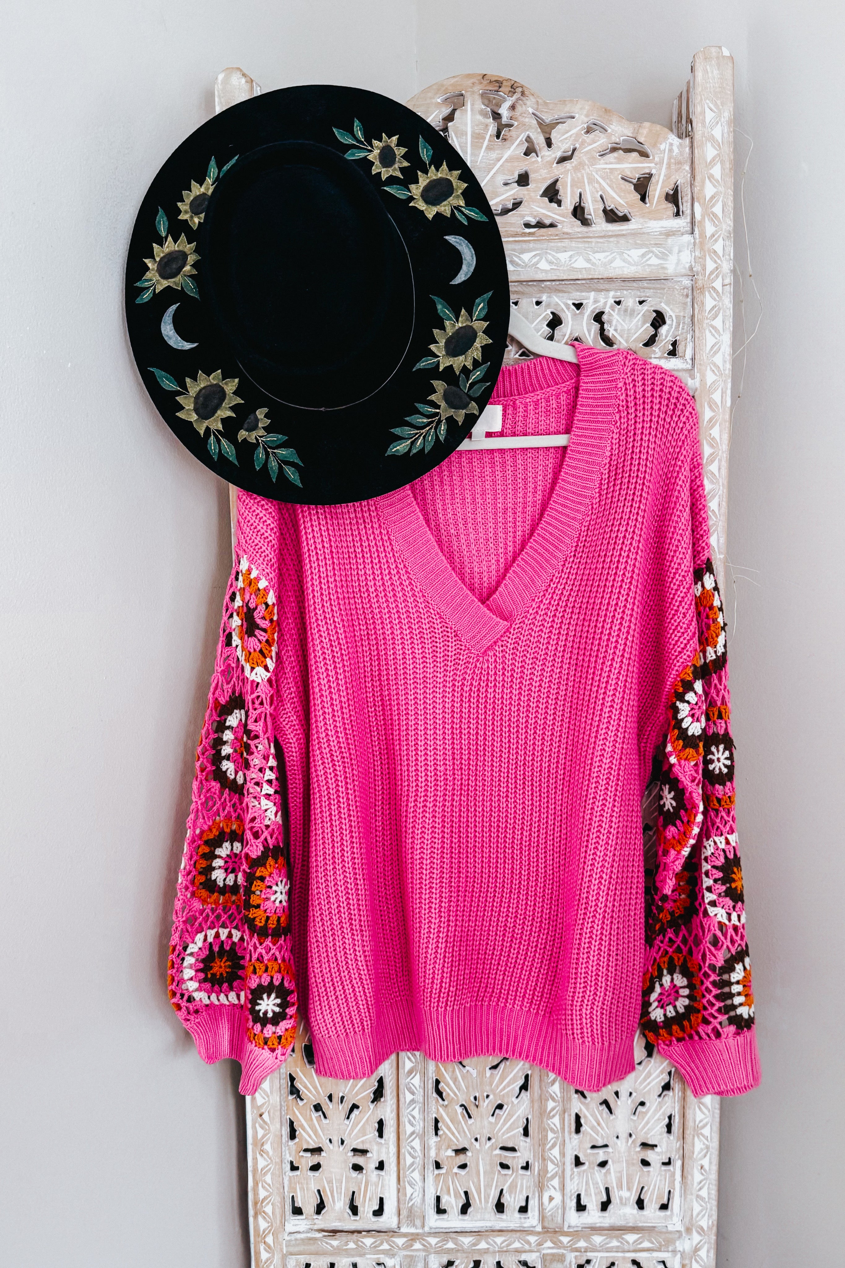Bright pink V-neck knit sweater with crochet patterned balloon sleeves hanging on ornate carved wooden panel with black hat above
