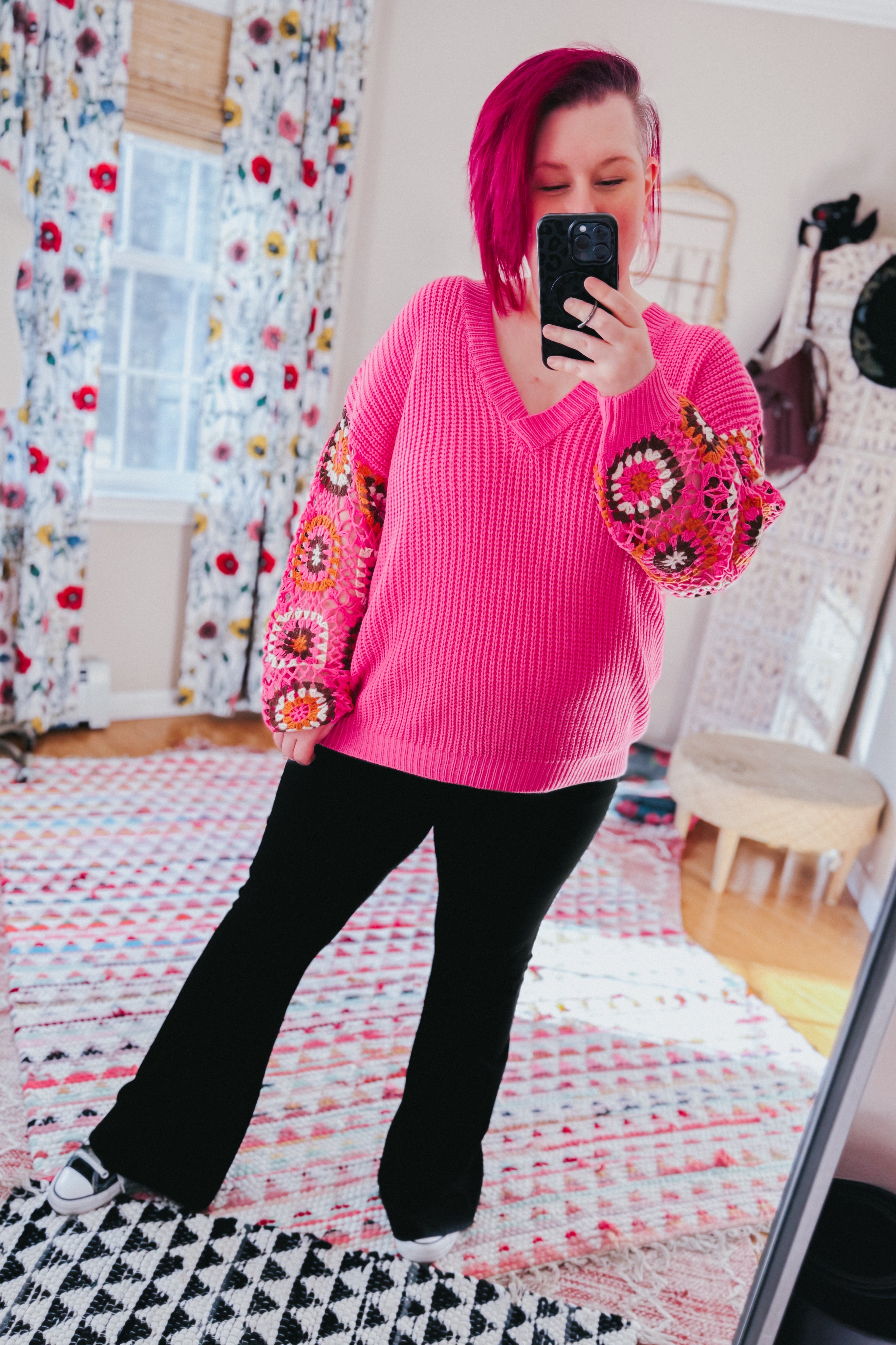Woman with short magenta hair wearing bright pink V-neck knit sweater with multicolored crochet sleeves and black pants indoors