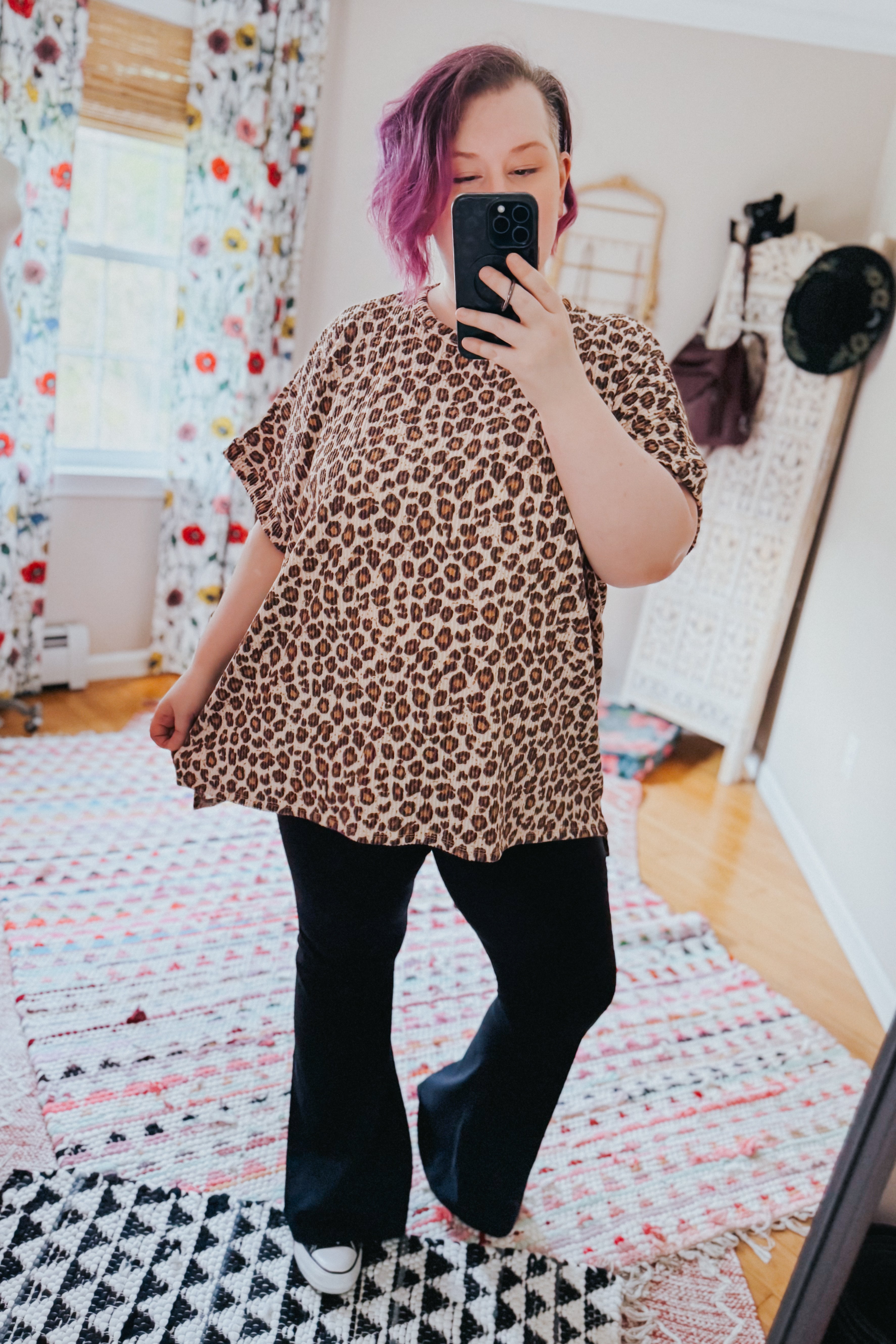 Woman taking mirror selfie wearing loose leopard print short-sleeve top with black flared pants and white sneakers indoors