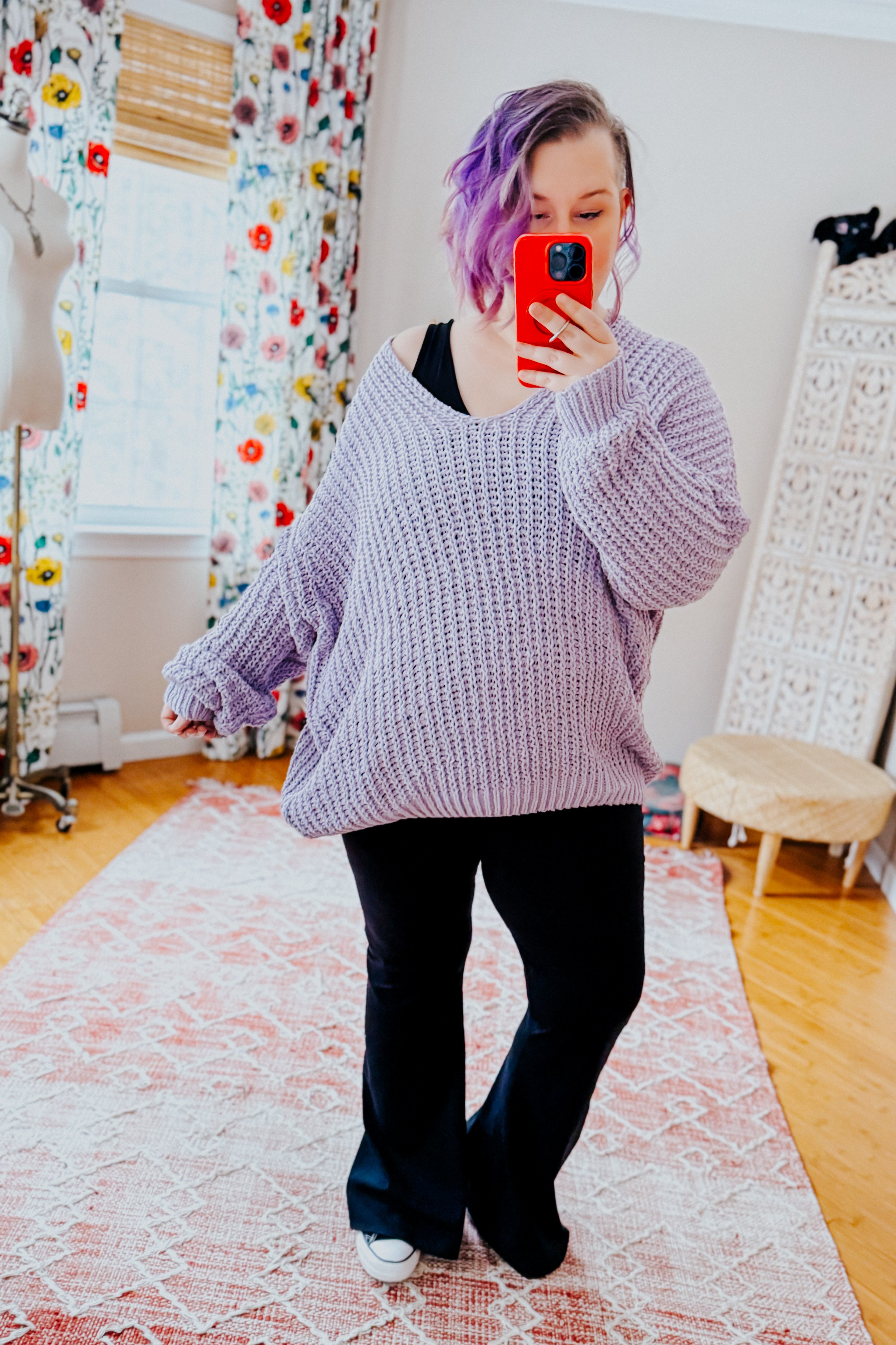 Woman with purple hair wearing oversized chunky lavender V-neck sweater and black flared pants taking selfie indoors