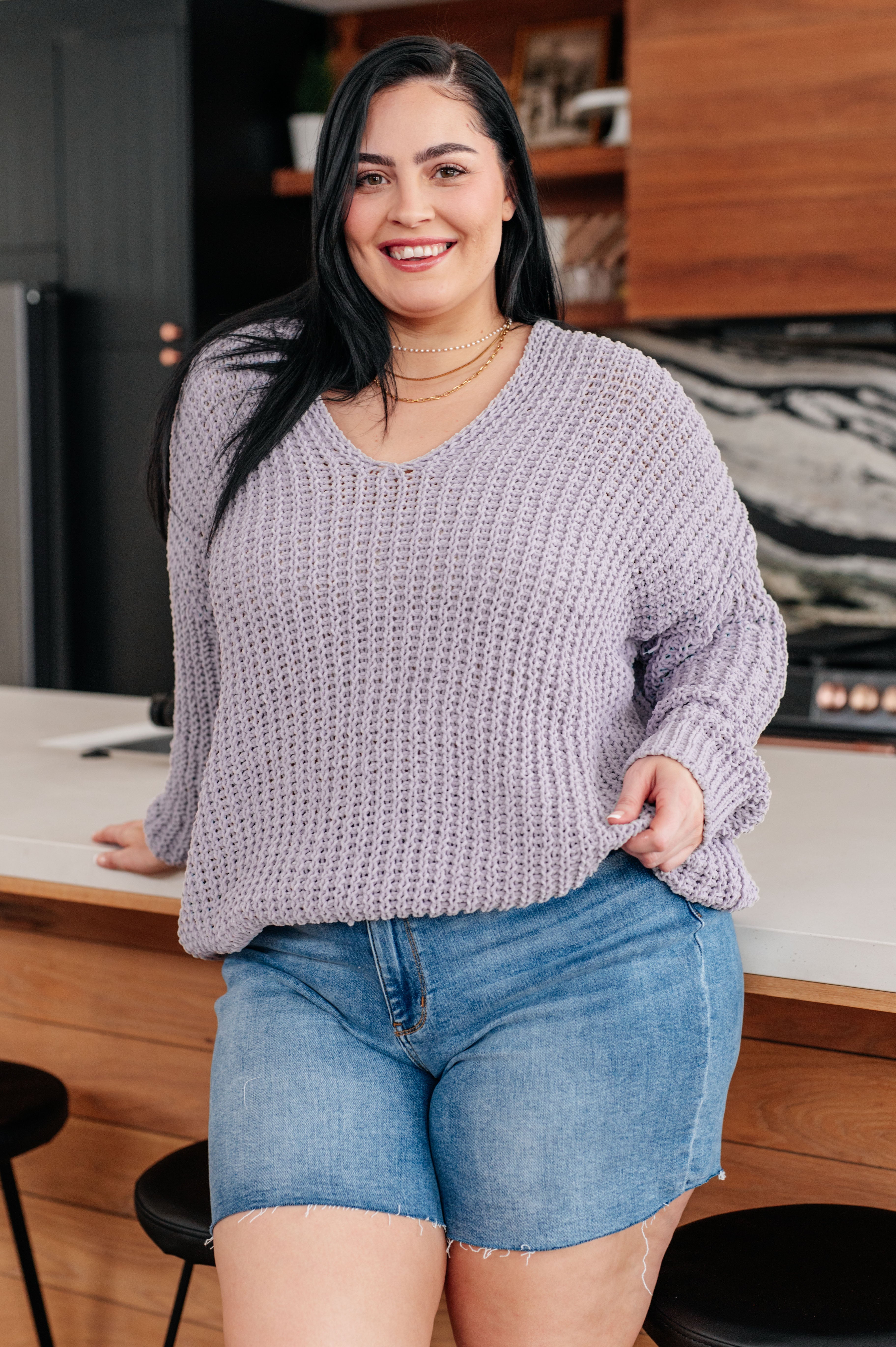 Smiling woman with long dark hair wearing a chunky lavender V-neck sweater and denim shorts indoors by kitchen island