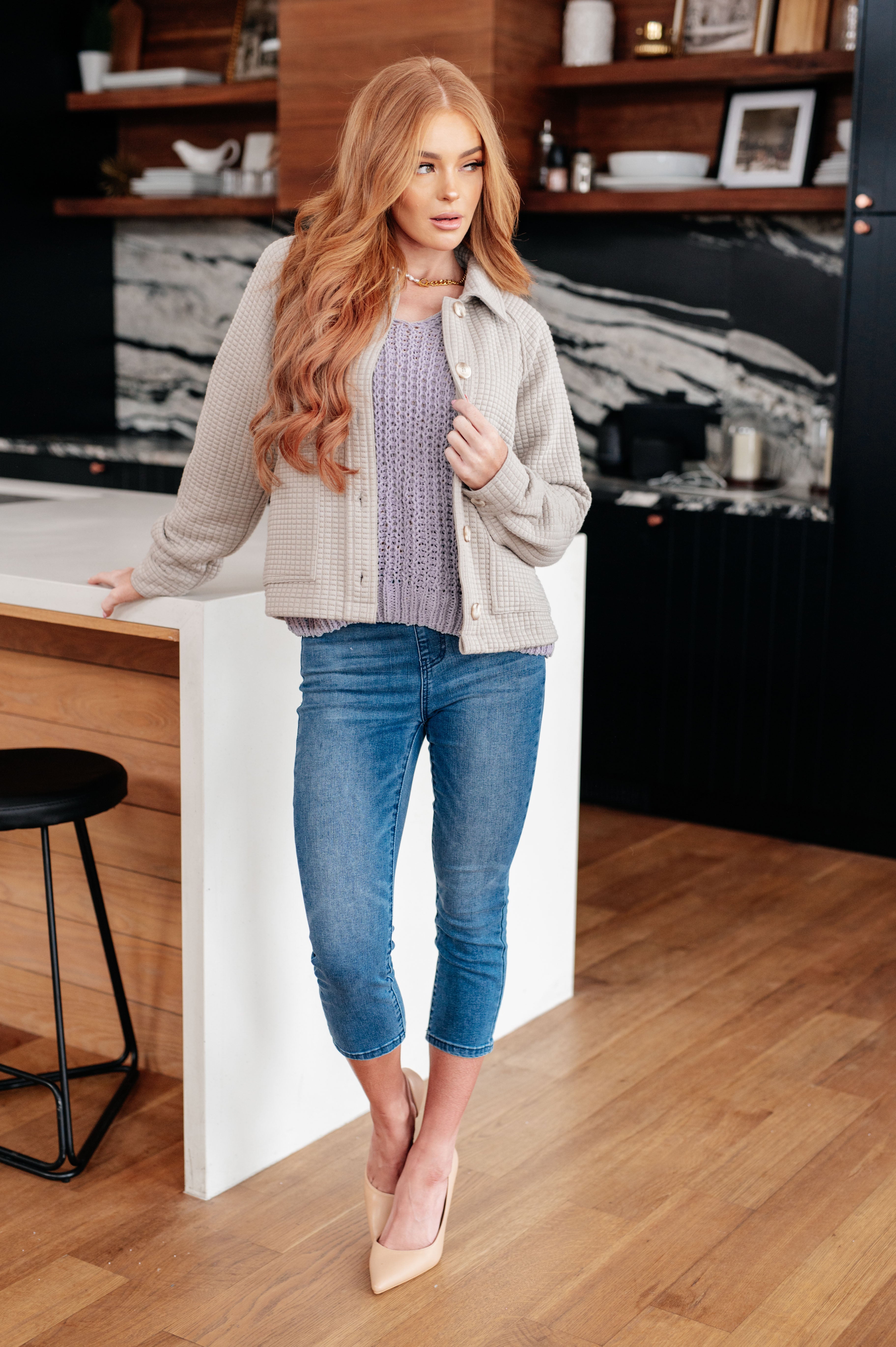 Red-haired woman wearing a lavender chunky V-neck sweater layered under a beige textured jacket with cropped jeans indoors