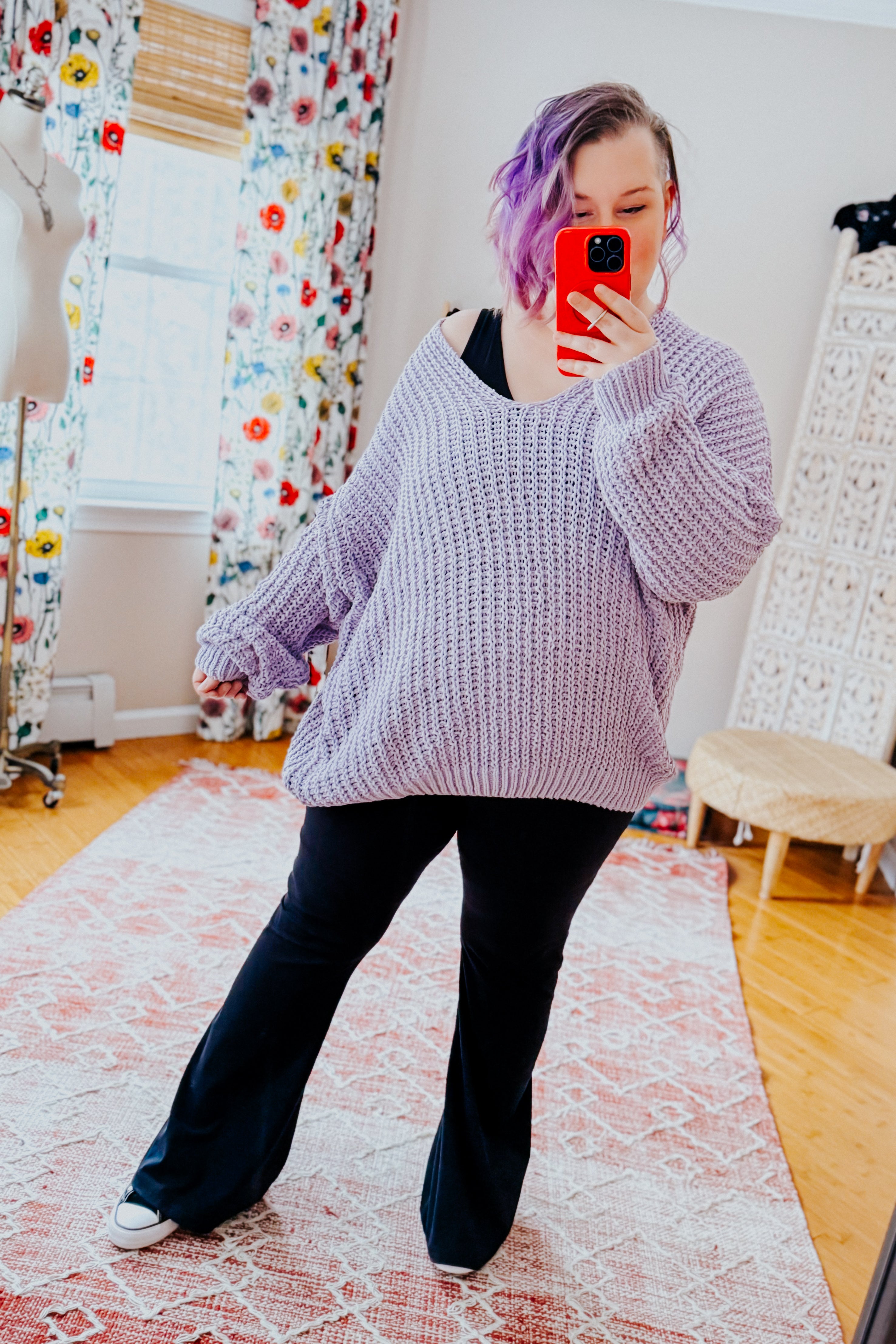 Woman with short purple hair wearing an oversized chunky lavender V-neck sweater and black pants taking selfie indoors