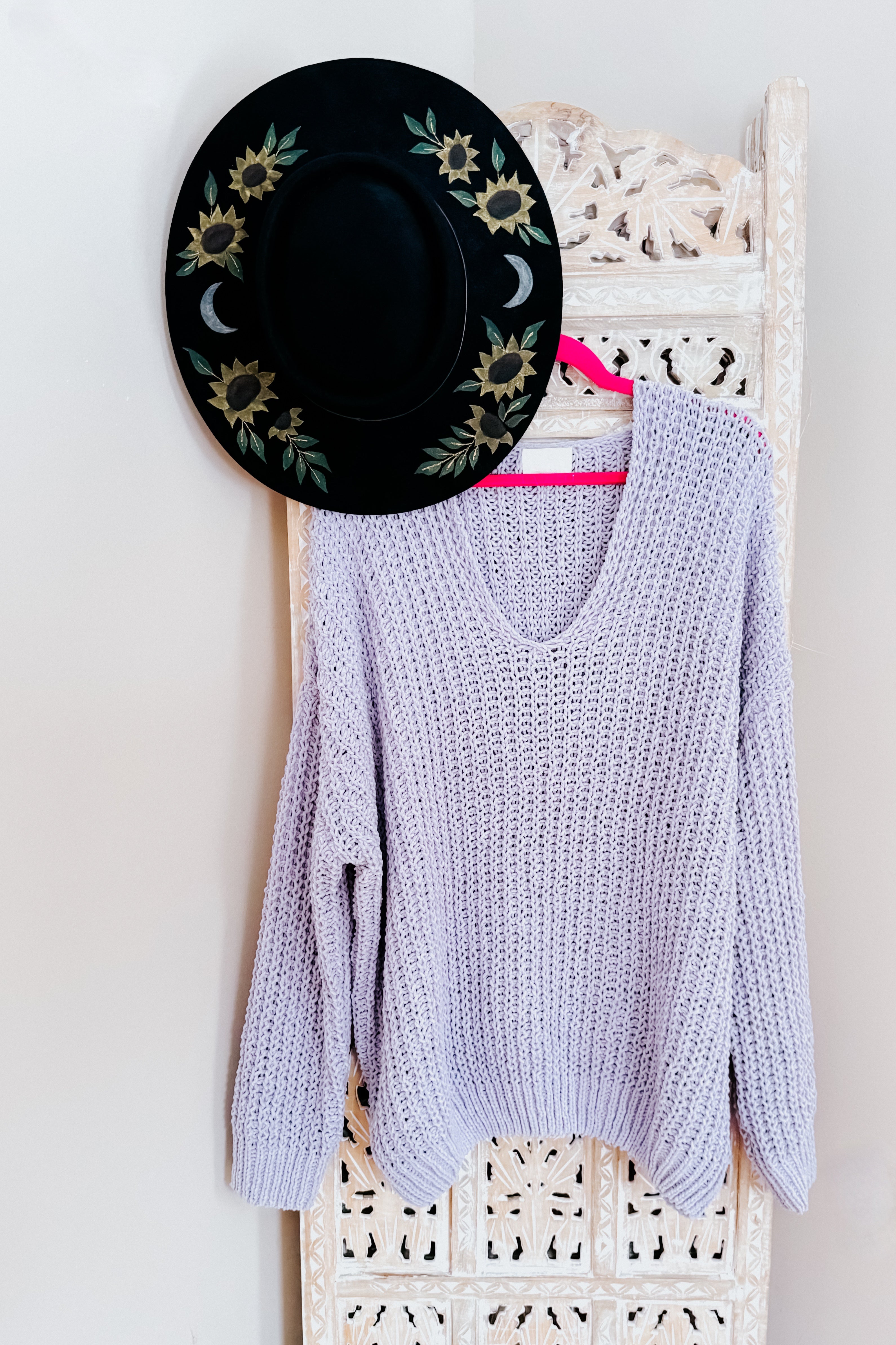 Chunky light purple V-neck sweater hanging on a pink hanger against a carved ivory decorative screen with a black floral hat