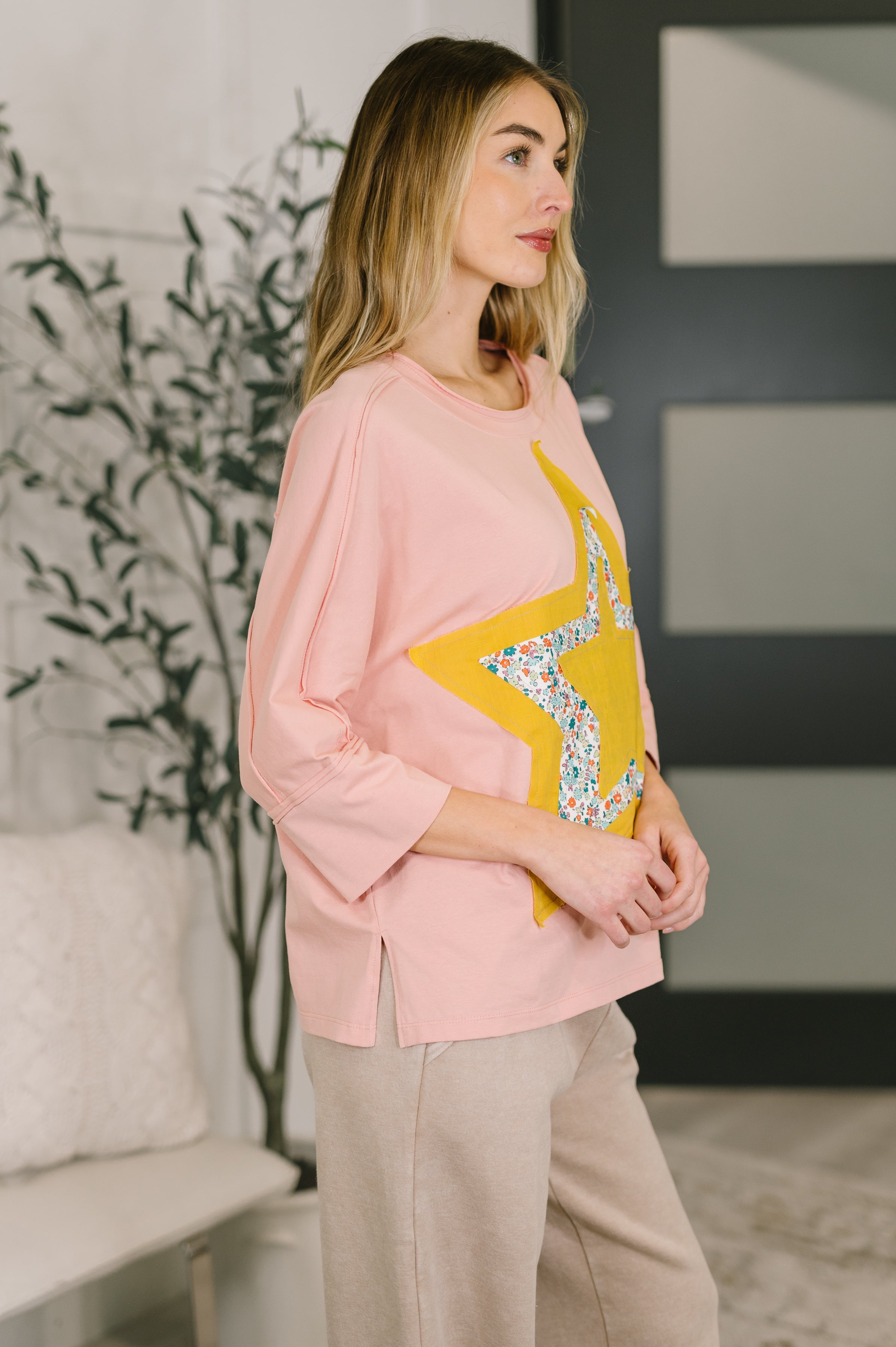 Woman wearing loose fit pink top with mixed print star patch detail and beige pants standing indoors side view