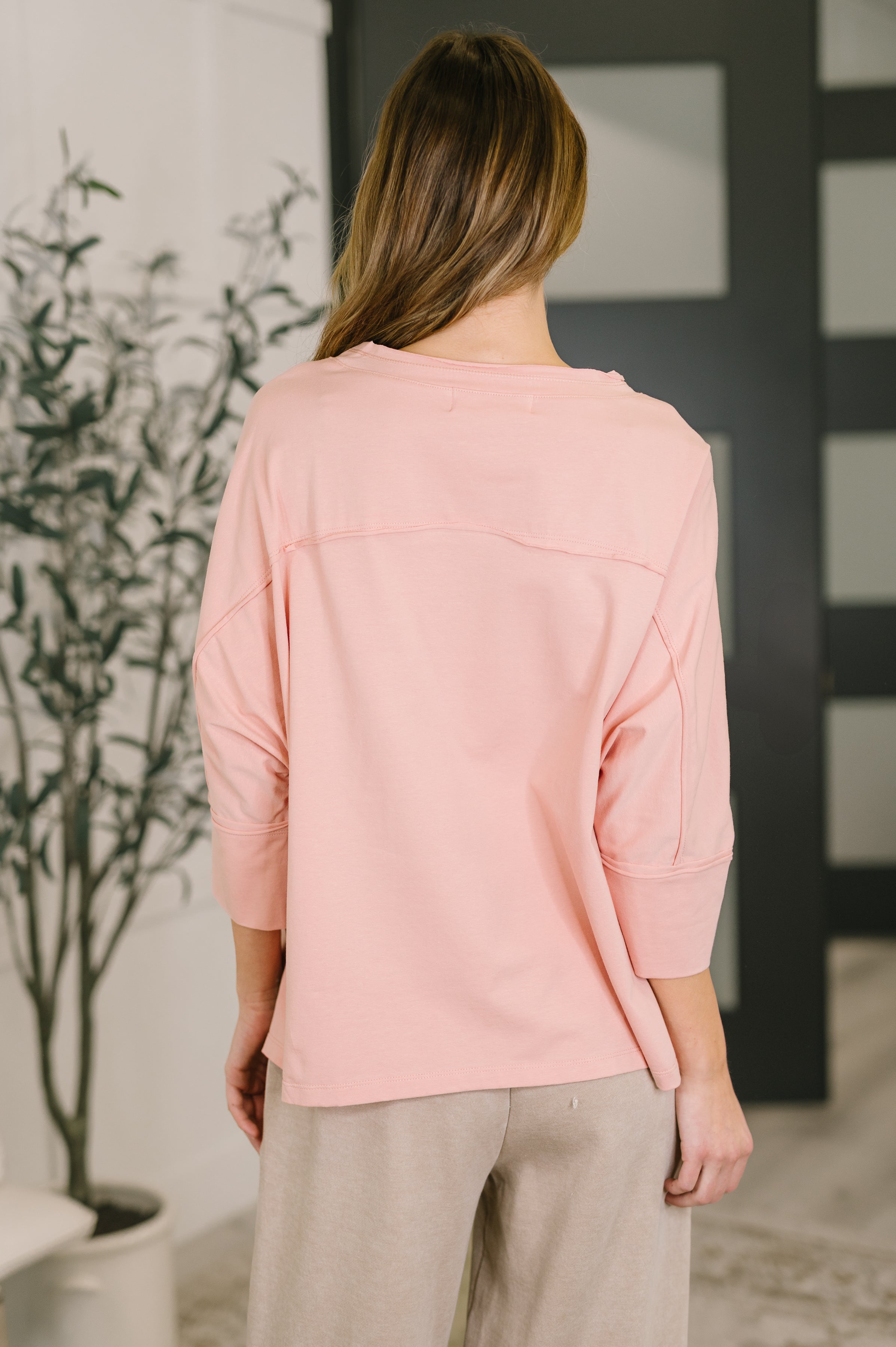 Woman wearing loose fit pastel pink top with three-quarter sleeves and beige pants indoors from back view