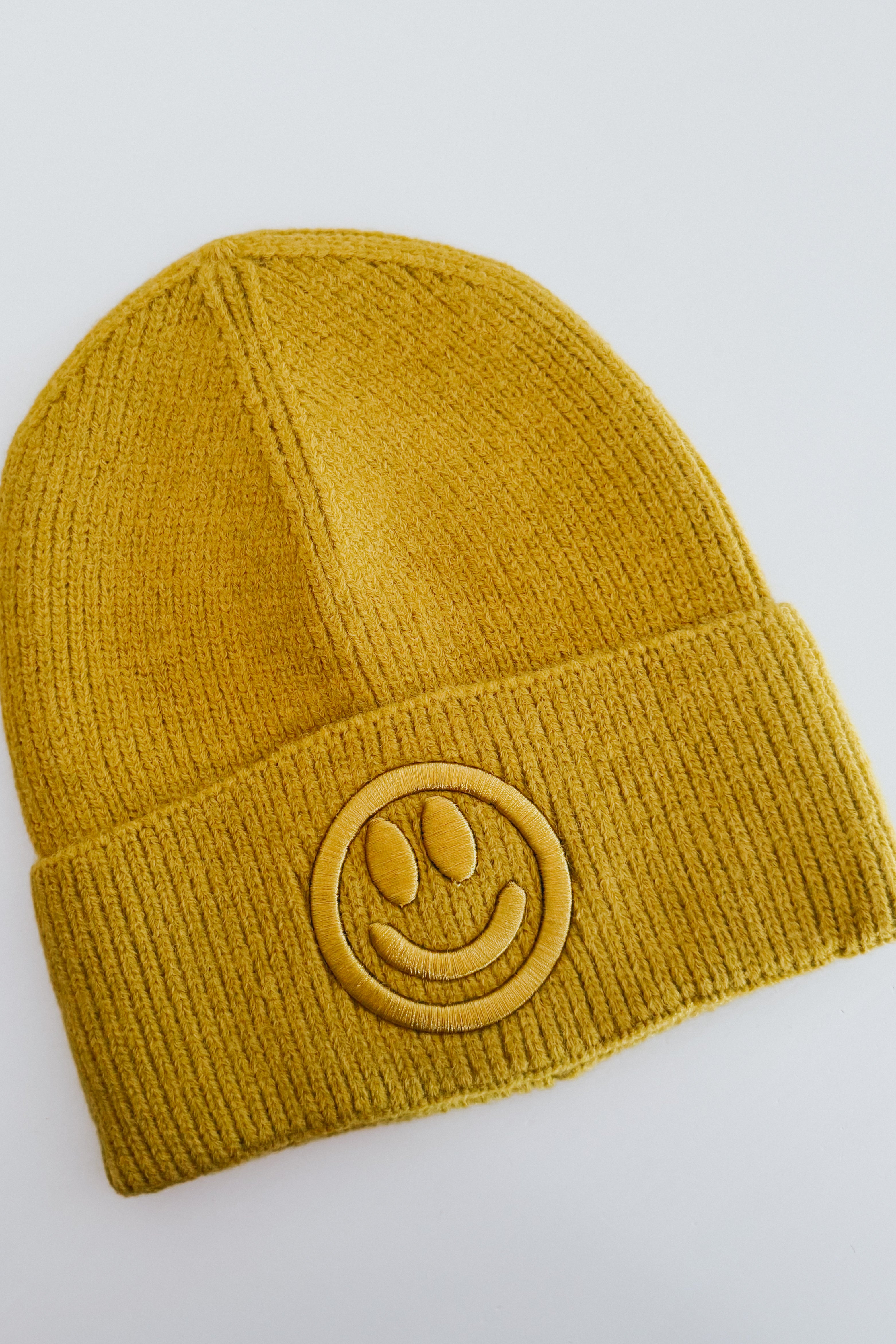 Mustard yellow ribbed knit beanie with embroidered smiley face patch on folded cuff against plain light background