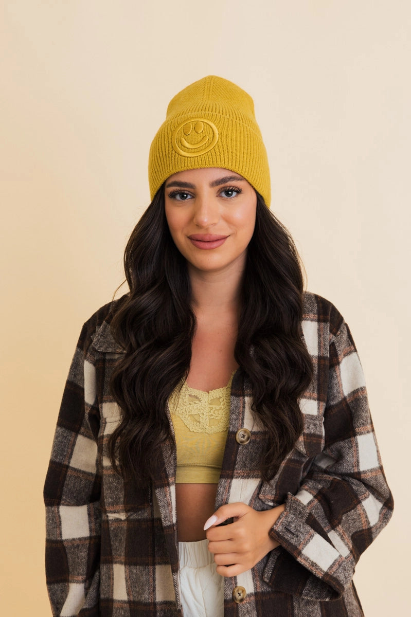 Smiling woman wearing mustard yellow ribbed knit beanie with embroidered smiley face and plaid jacket on beige background