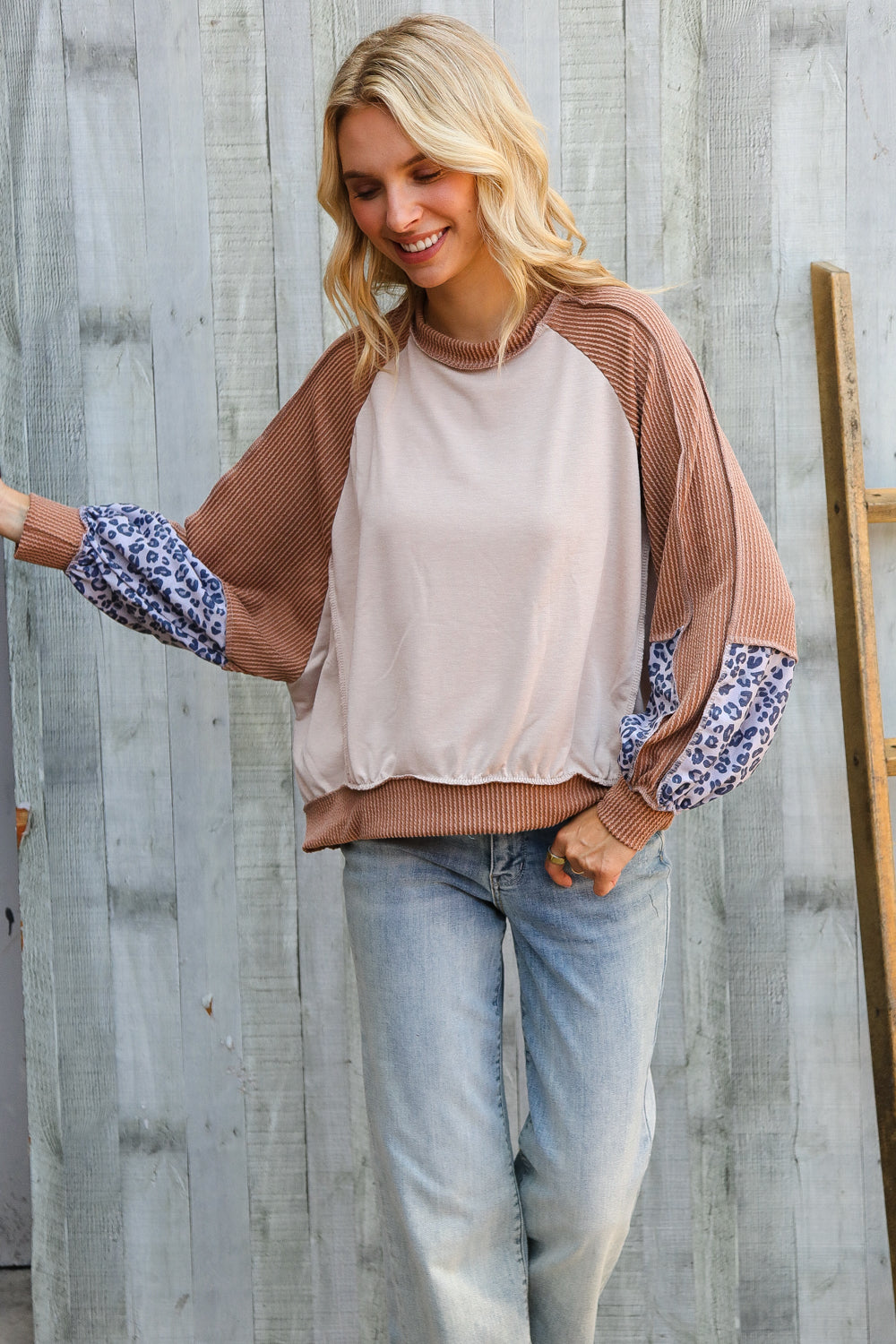 Woman wearing beige and brown dolman colorblock terry top with leopard print sleeves and light wash jeans standing by wooden wall