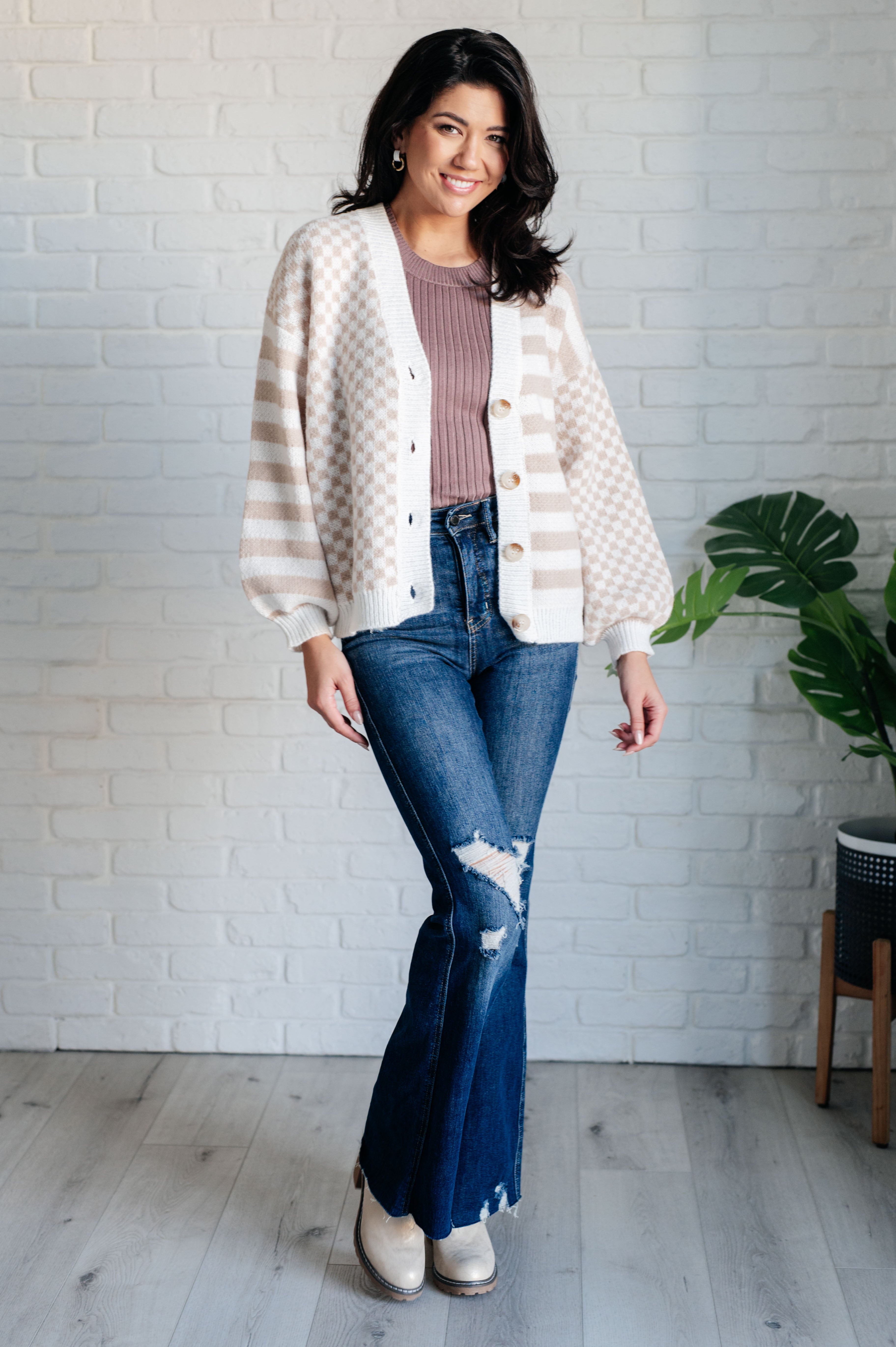 Woman wearing beige and white checkered and striped cardigan with button front, mauve ribbed top, and distressed flared jeans