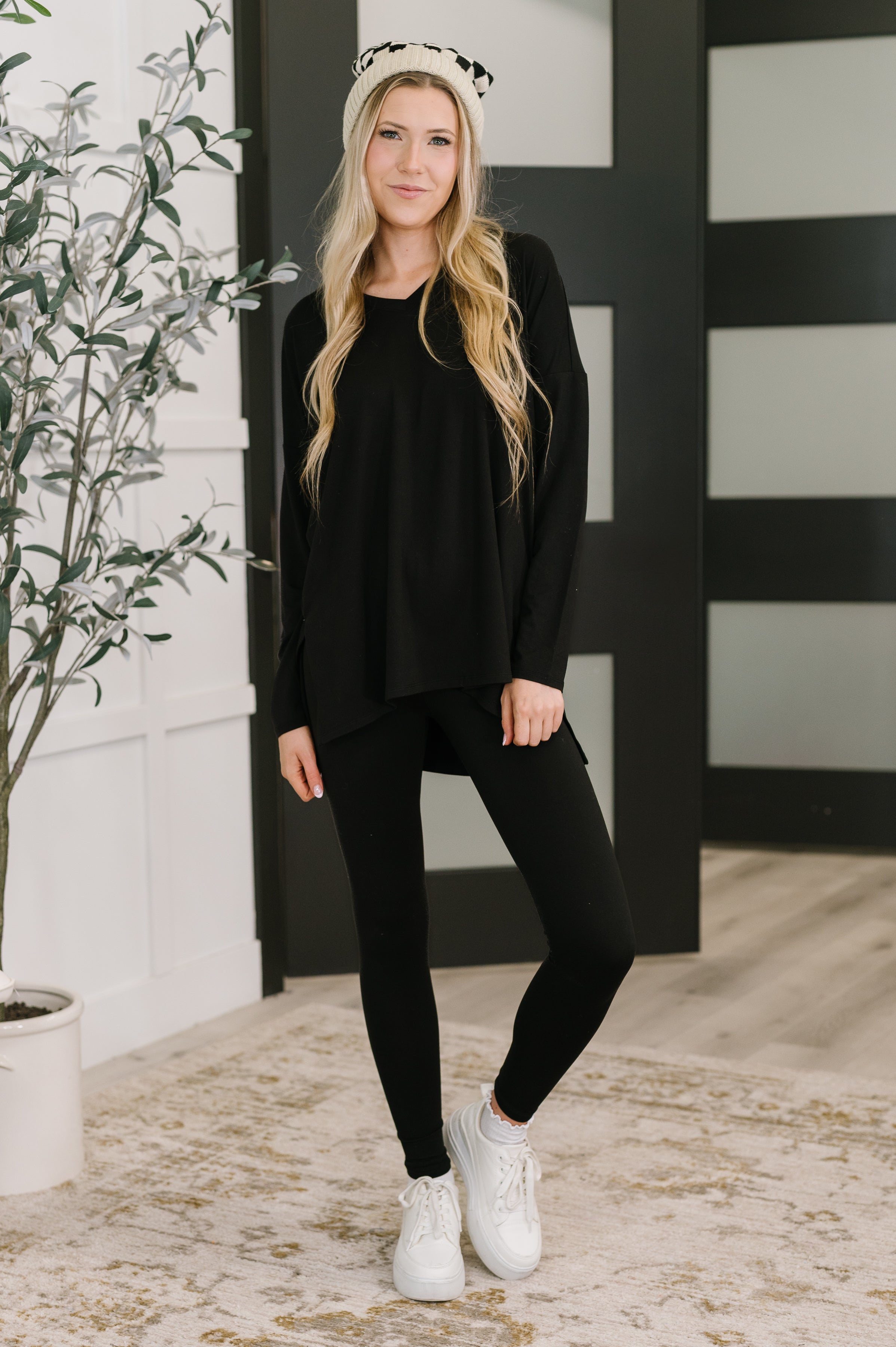 Blonde woman wearing bone-colored checkered beanie with loose black top, black leggings, and white sneakers indoors