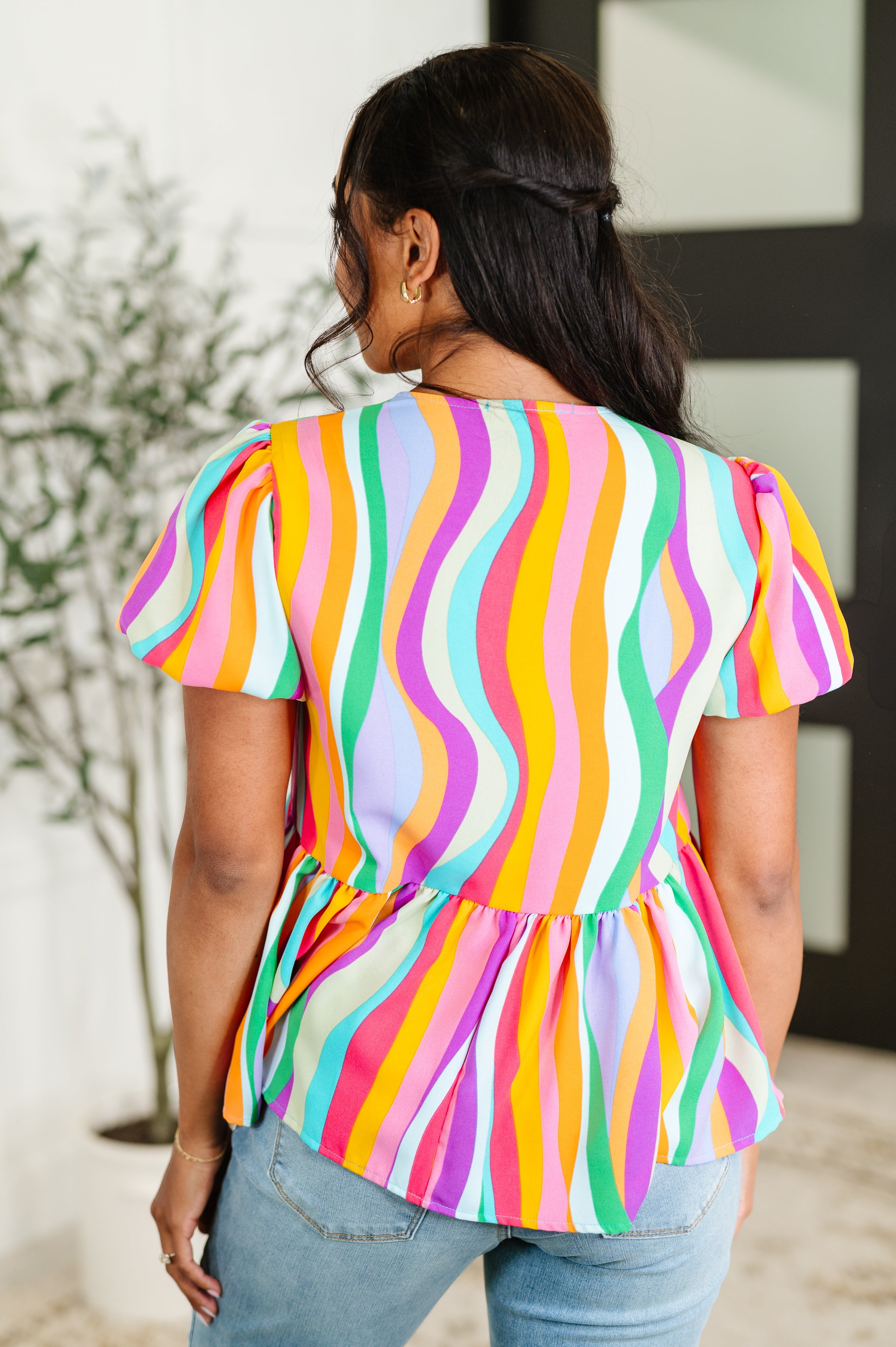 Woman wearing colorful wavy striped puff sleeve blouse in mint, orange, purple, and pink, paired with light blue jeans
