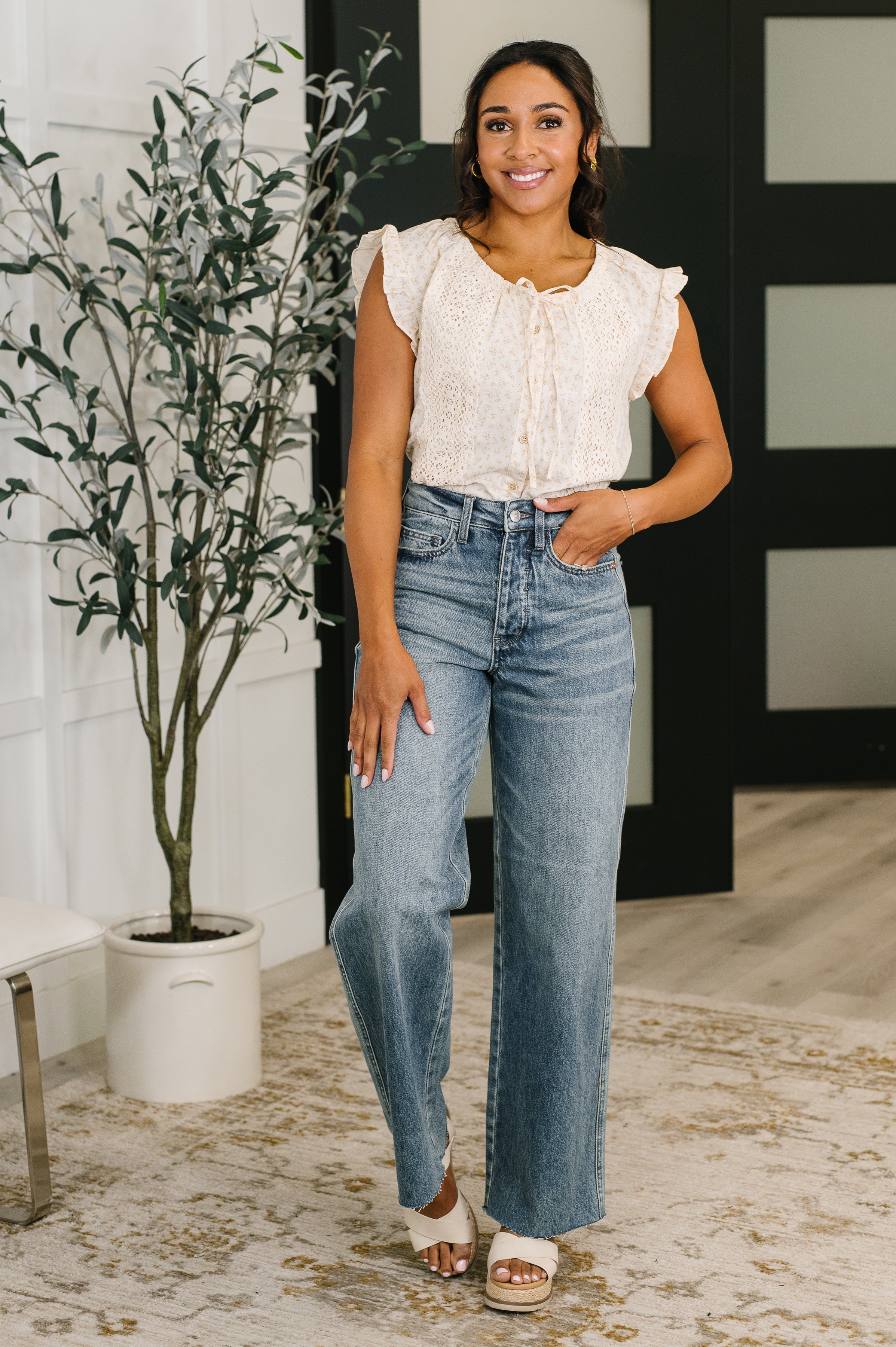 Woman wearing blue high waist baggy jeans and cream ruffled sleeveless blouse standing indoors near plant and chair