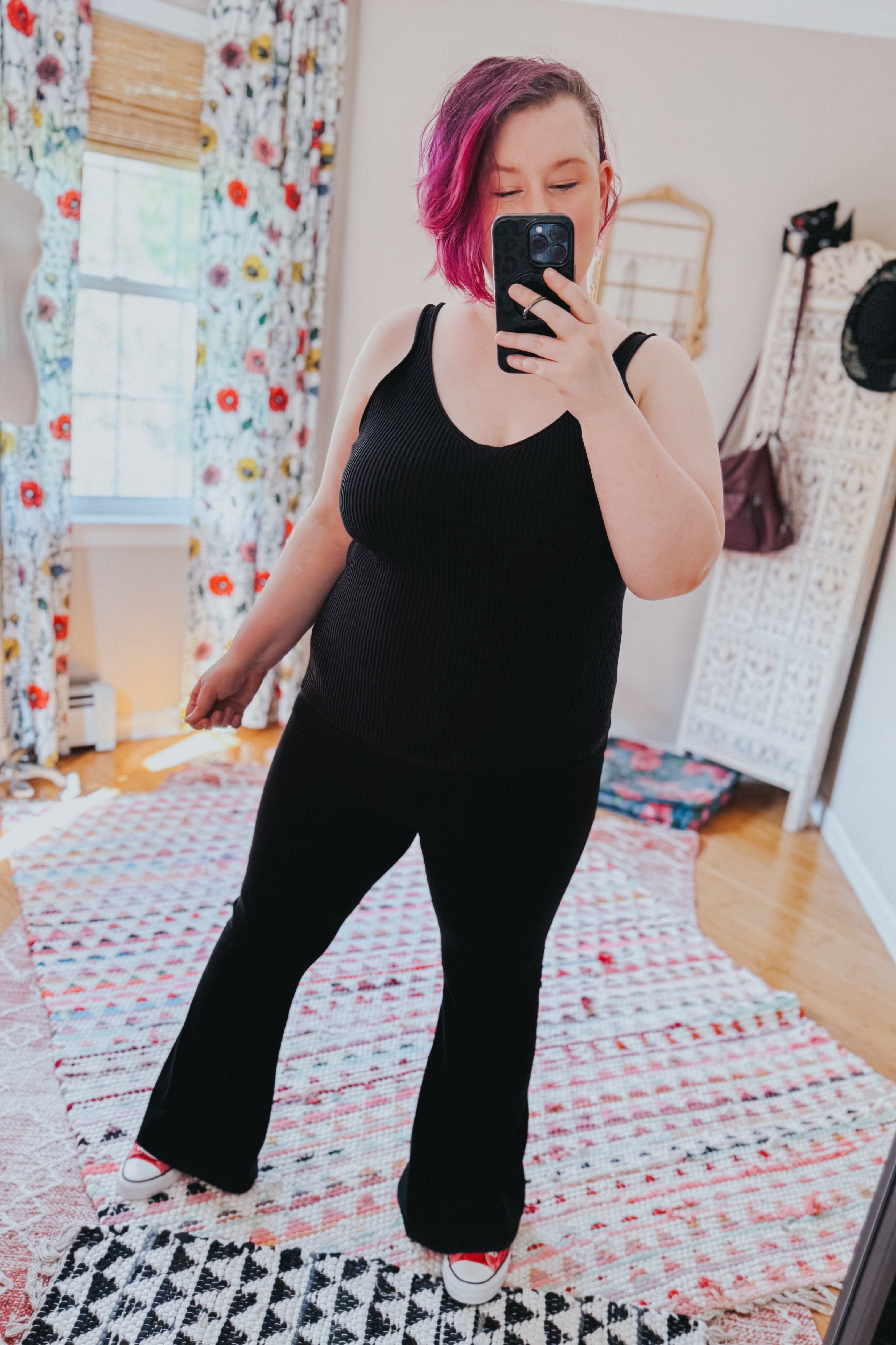 Woman with short pink hair wearing a black ribbed tank top and black flared pants taking a mirror selfie indoors