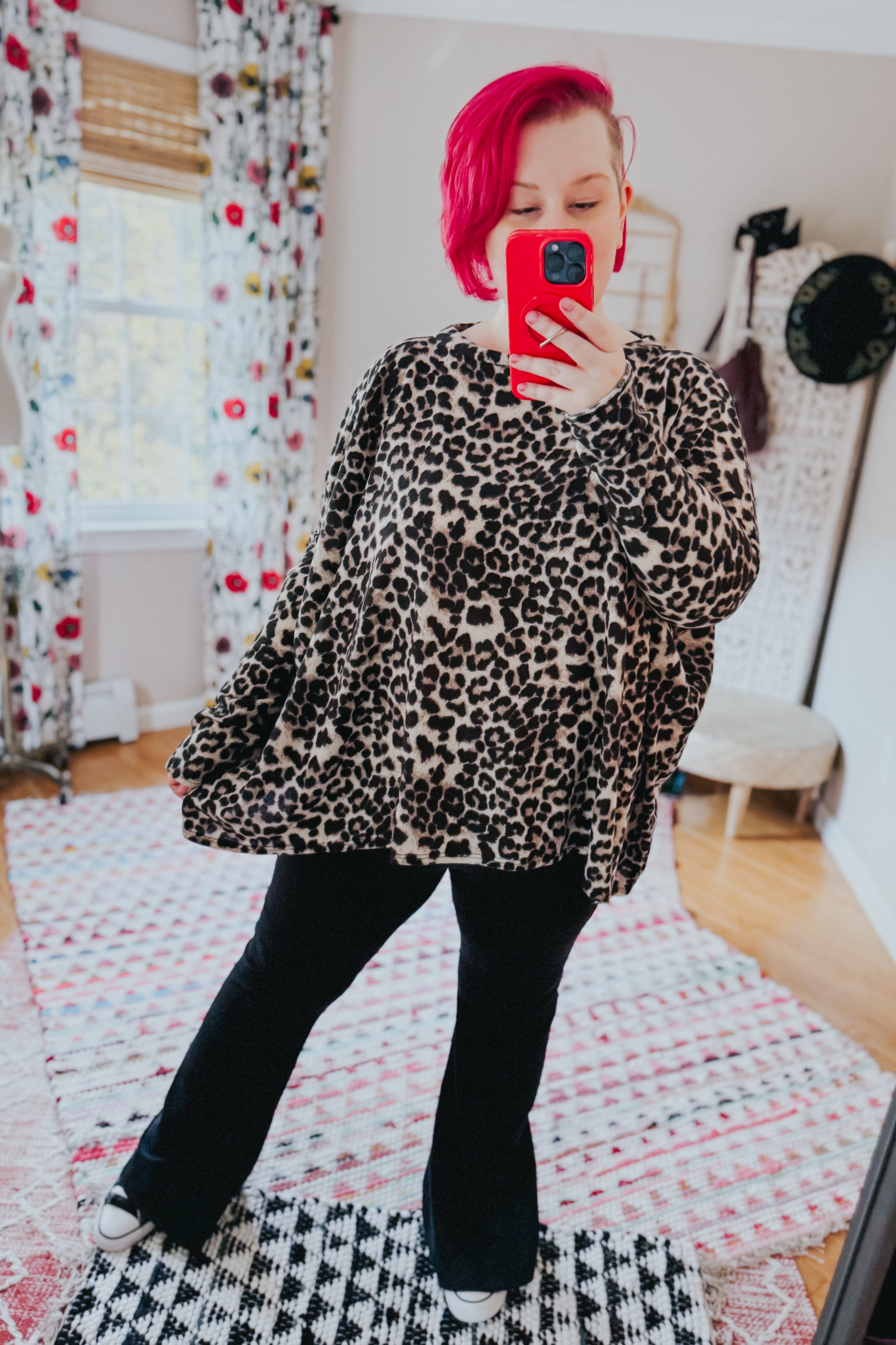 Woman with bright pink hair wearing oversized leopard print long sleeve top and black pants taking mirror selfie indoors