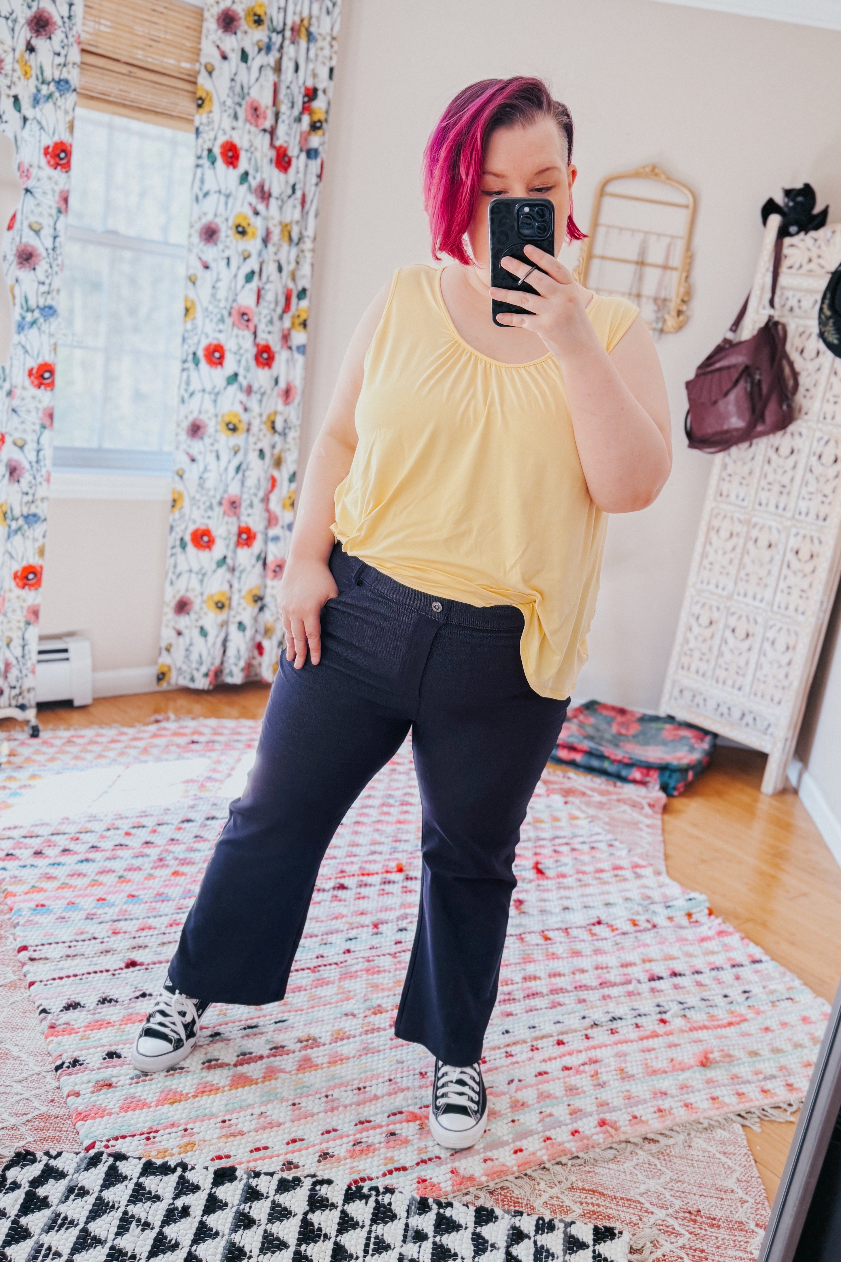 Woman with short pink hair wearing sleeveless yellow top and Classic Charm Cropped Pants taking indoor selfie in colorful room