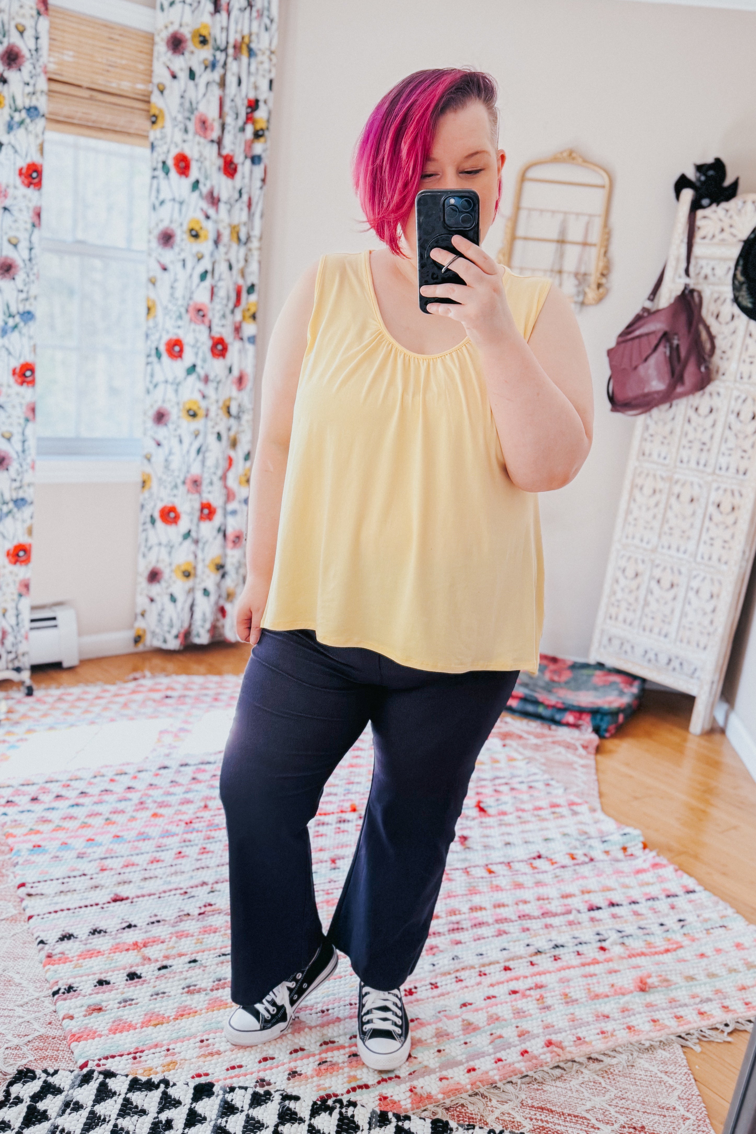 Woman with pink hair wearing sleeveless yellow top and Classic Charm Cropped Pants taking indoor mirror selfie in colorful room