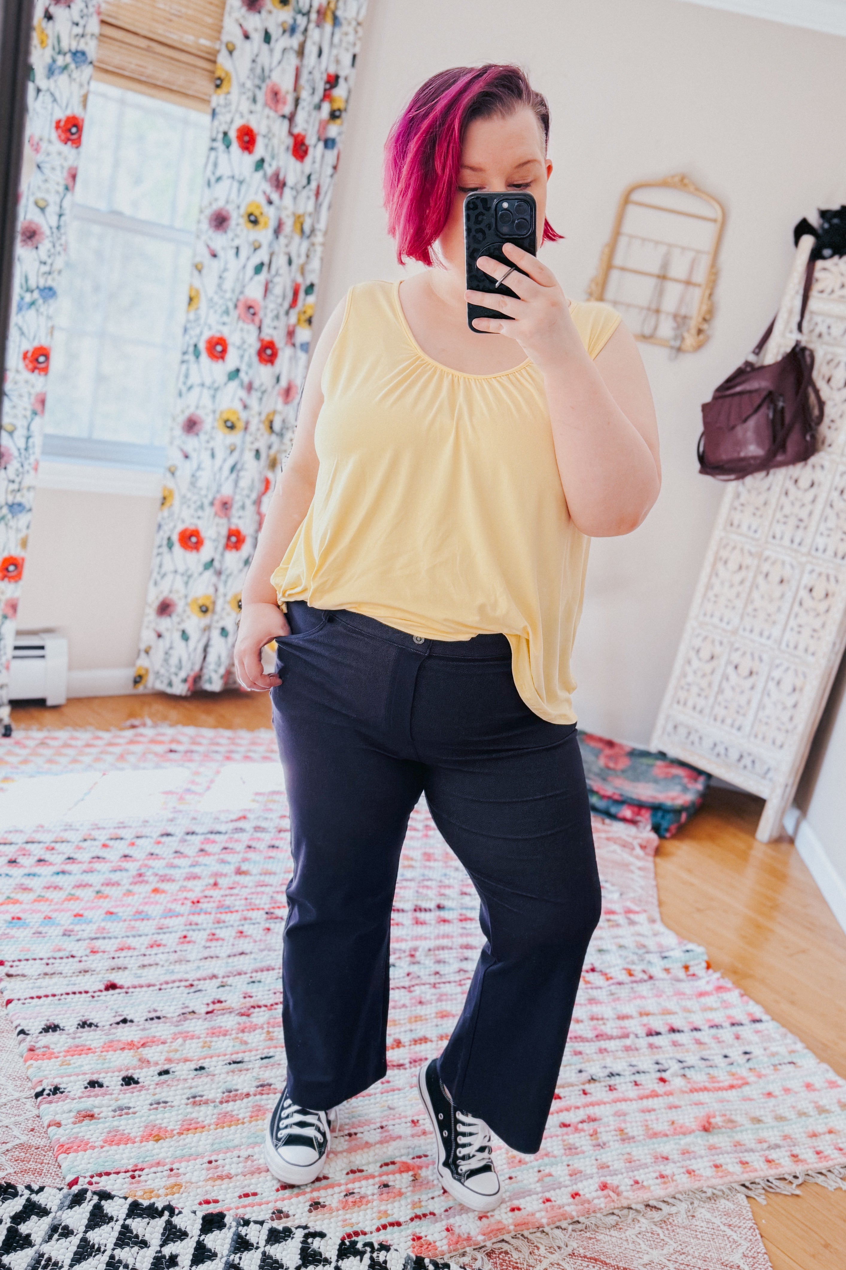 Woman with short pink hair wearing sleeveless yellow top and black cropped pants taking mirror selfie indoors