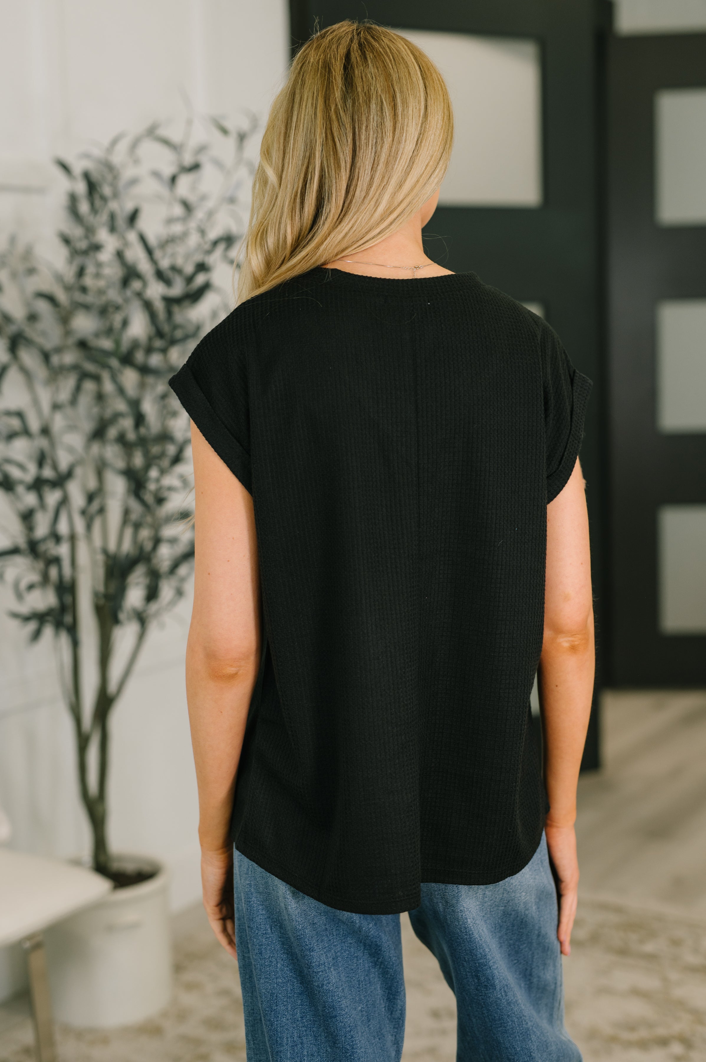 Back view of woman in relaxed black short-sleeve top paired with casual blue jeans indoors near plant and door